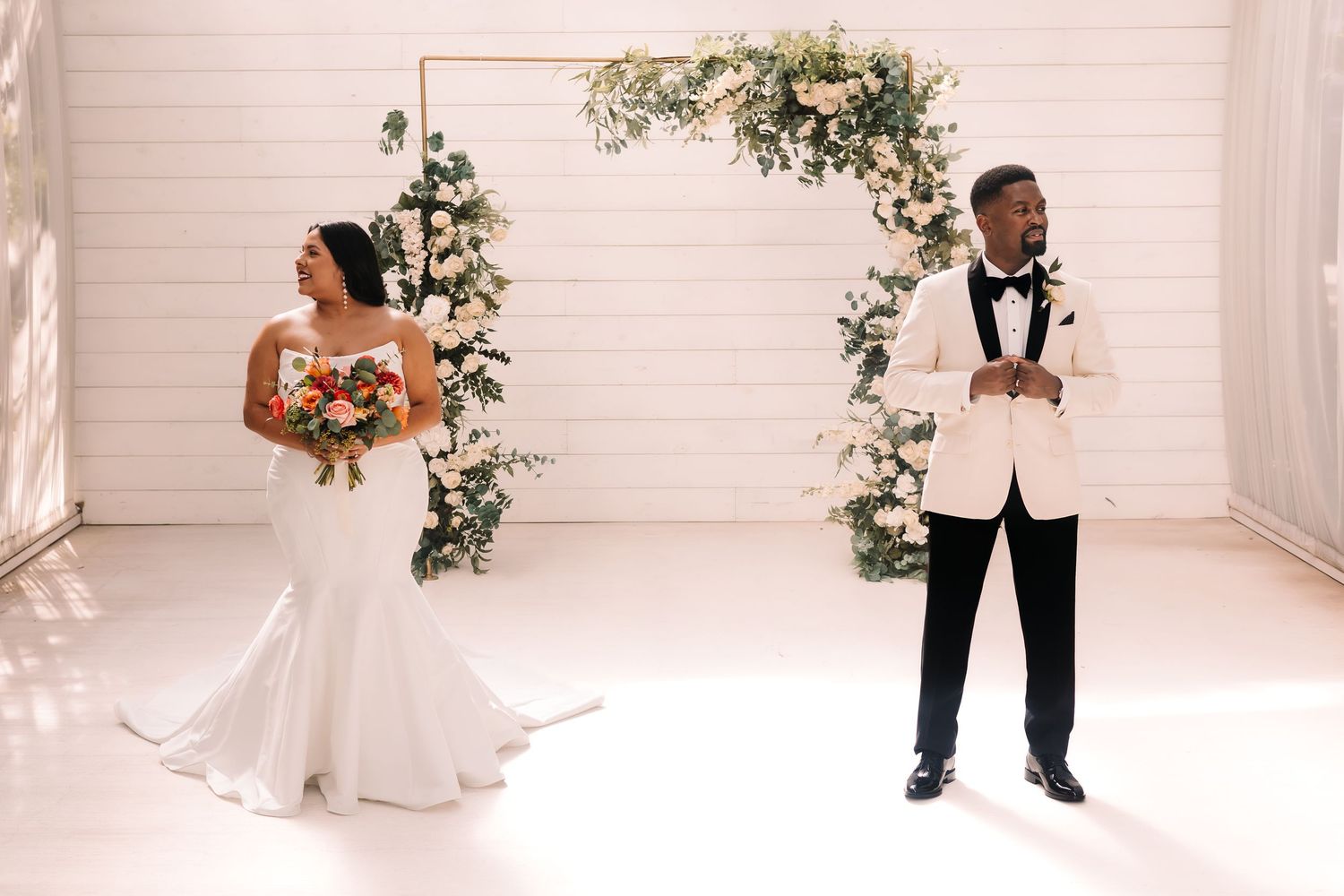 Bride in mermaid gown and groom in white tuxedo standing before floral arch in white venue in Houston Texas.