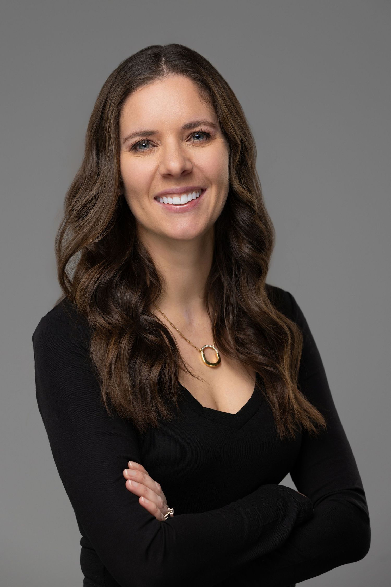 Professional headshot of a smiling woman with long brown hair wearing a black sweater and necklace against a gray background.