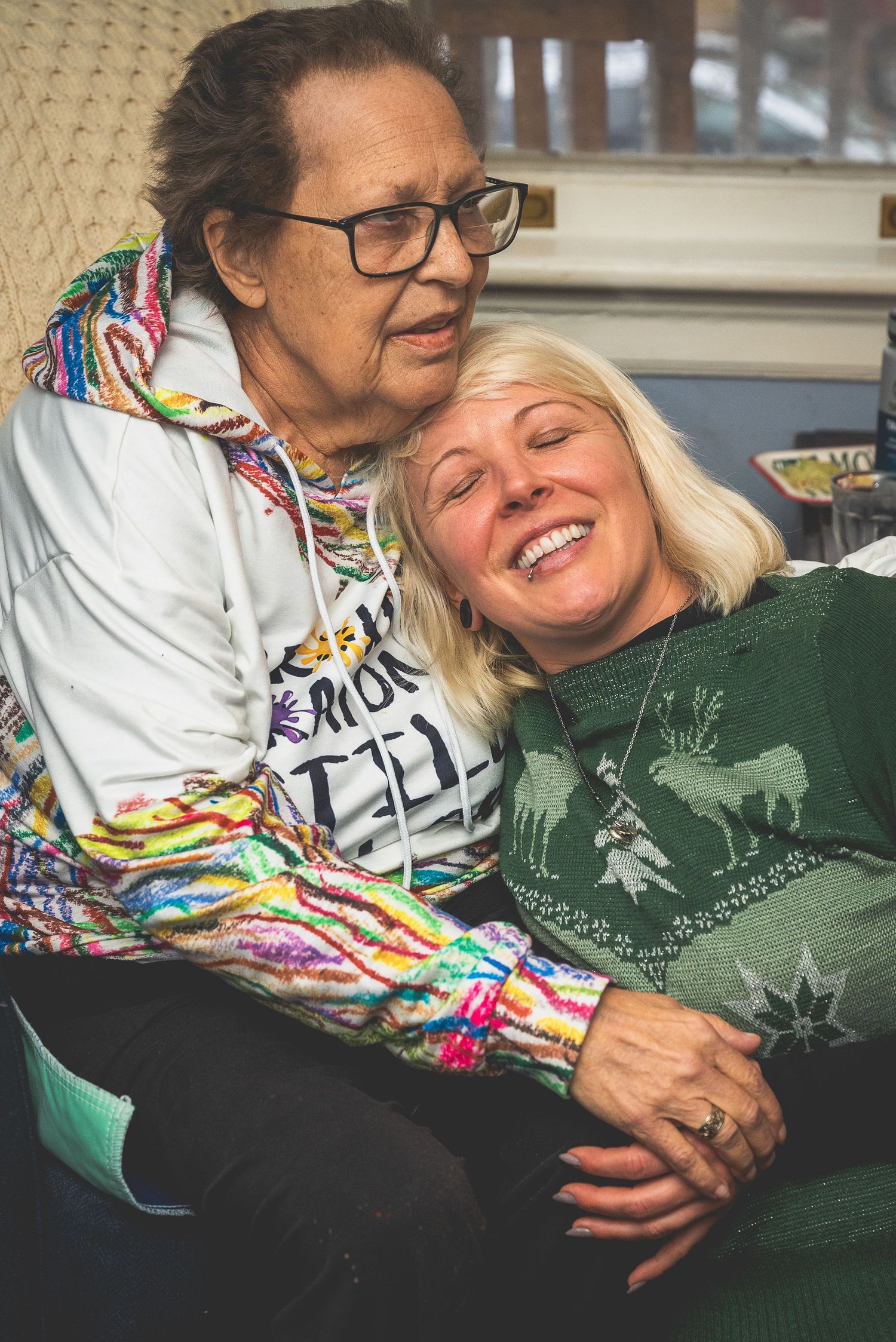 Two women, one with glasses and a colorful scarf, embrace and smile warmly together.