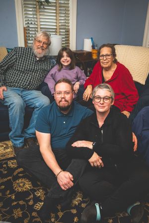 A group of five people, including an older man with a beard, a younger woman, and three other adults, are sitting together on a couch.