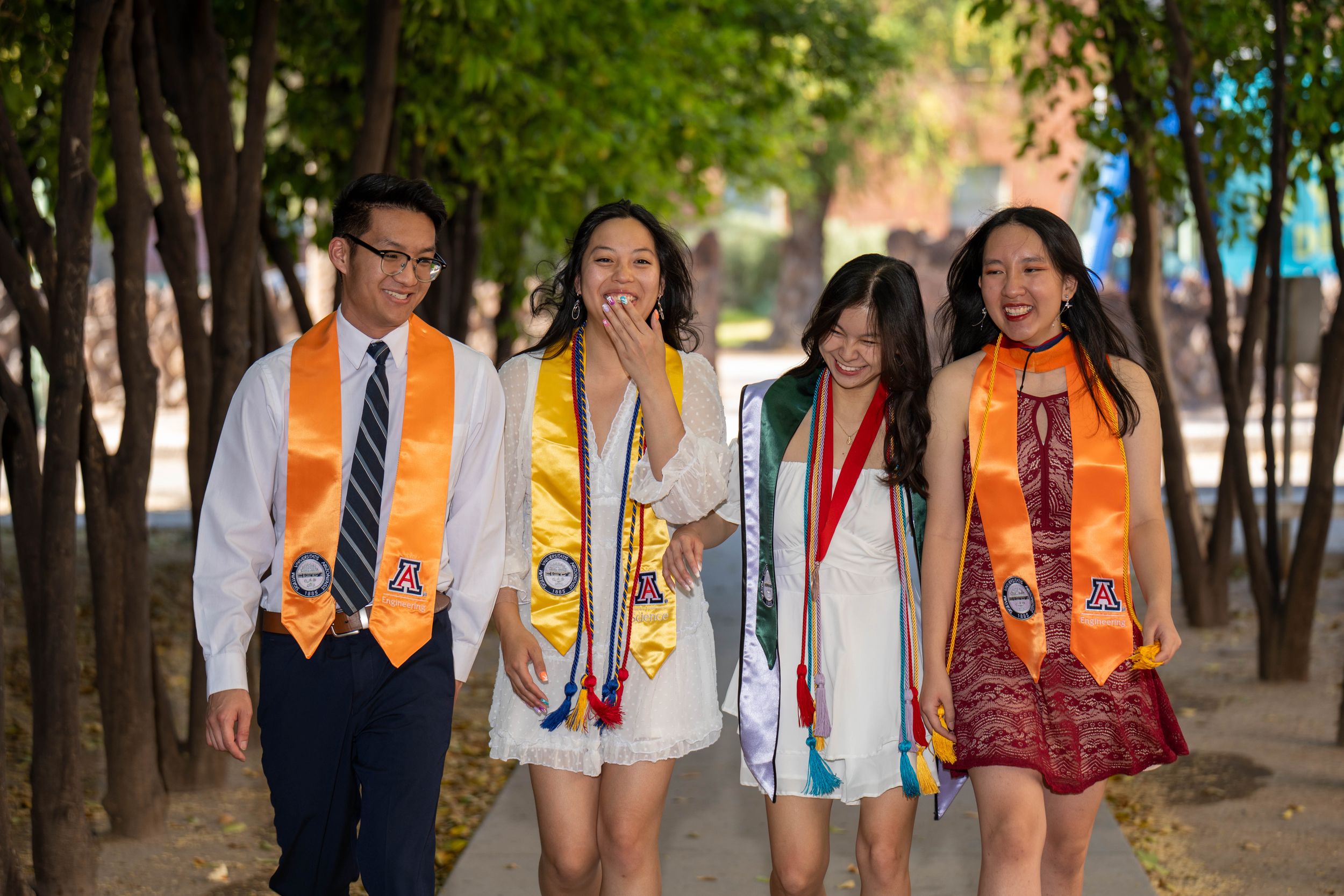 Candid photo of four U of A graduation students in stoles and cords walking and laughing together on university grounds.