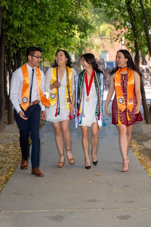 Group of four University of Arizona graduates in caps, gowns, and honors cords walking candidly down orange grove on the University Campus.