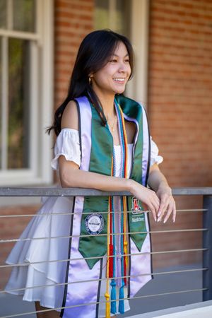 Grad student in cap, gown, and stoles leaning against the Old Main railing on the University of Arizona campus by N&D Media.