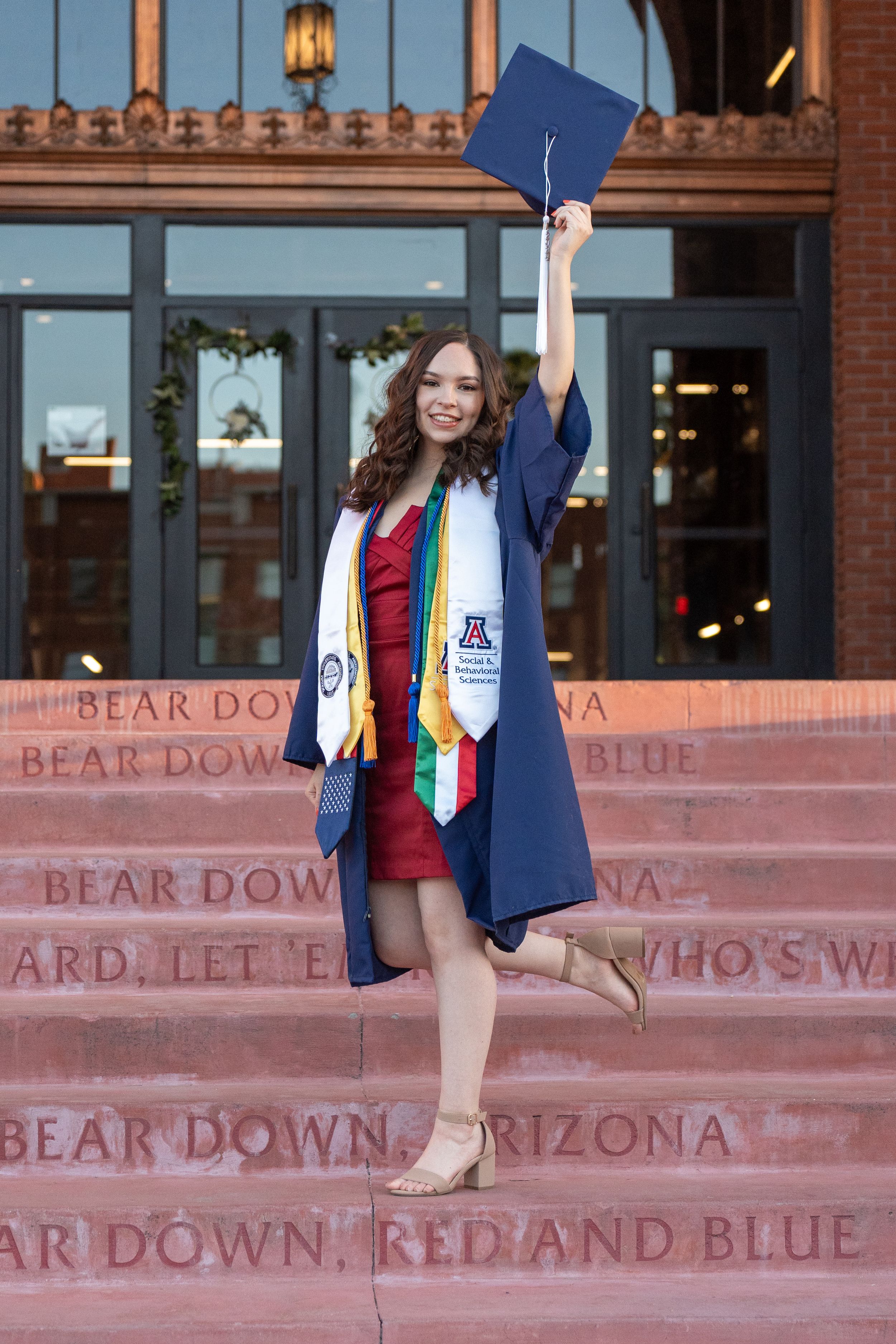 Female UofA graduate raising her cap on the engraved library steps in a victorious pose, captured by N&D Media Production.