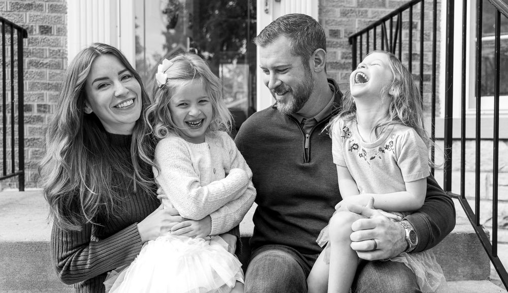 A black and white photograph of a family of four, including a mother, father, and two young daughters, sitting together outdoors.