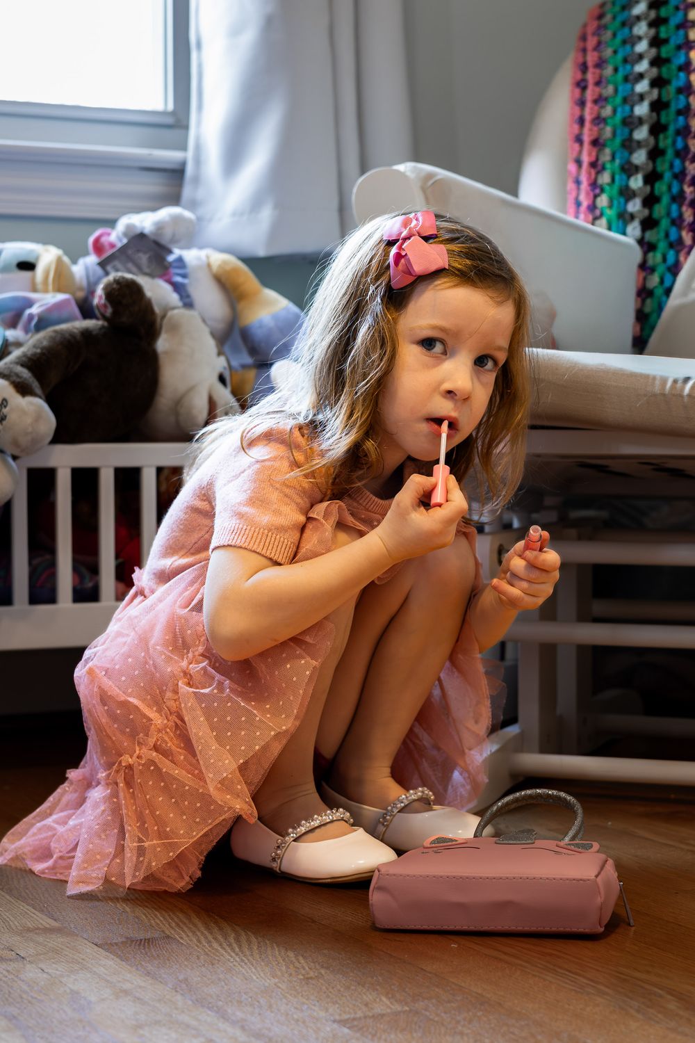 A young girl wearing a pink dress sits on the floor playing with toys in a cozy bedroom setting.