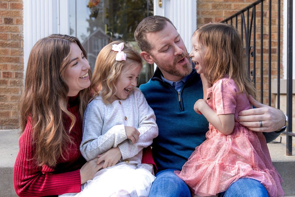 A happy family of four, including a father, mother, and two young daughters, sitting together outdoors laughing.