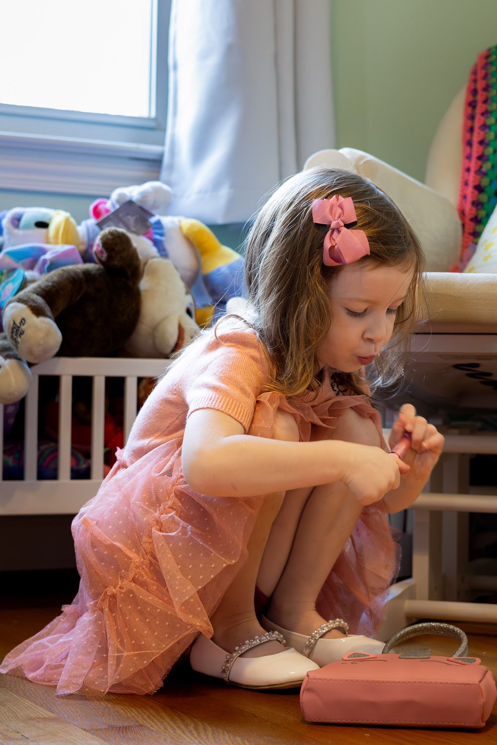 A young girl in a pink tulle dress sits on the floor playing with toys in a bedroom.