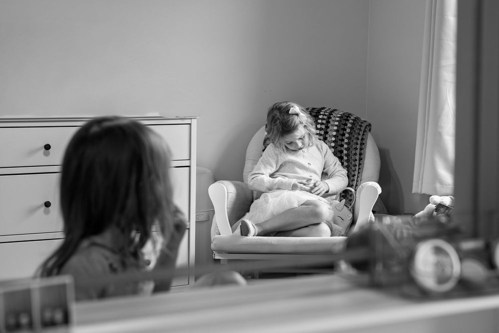 A black and white image shows a child sitting in a chair, playing in her room.