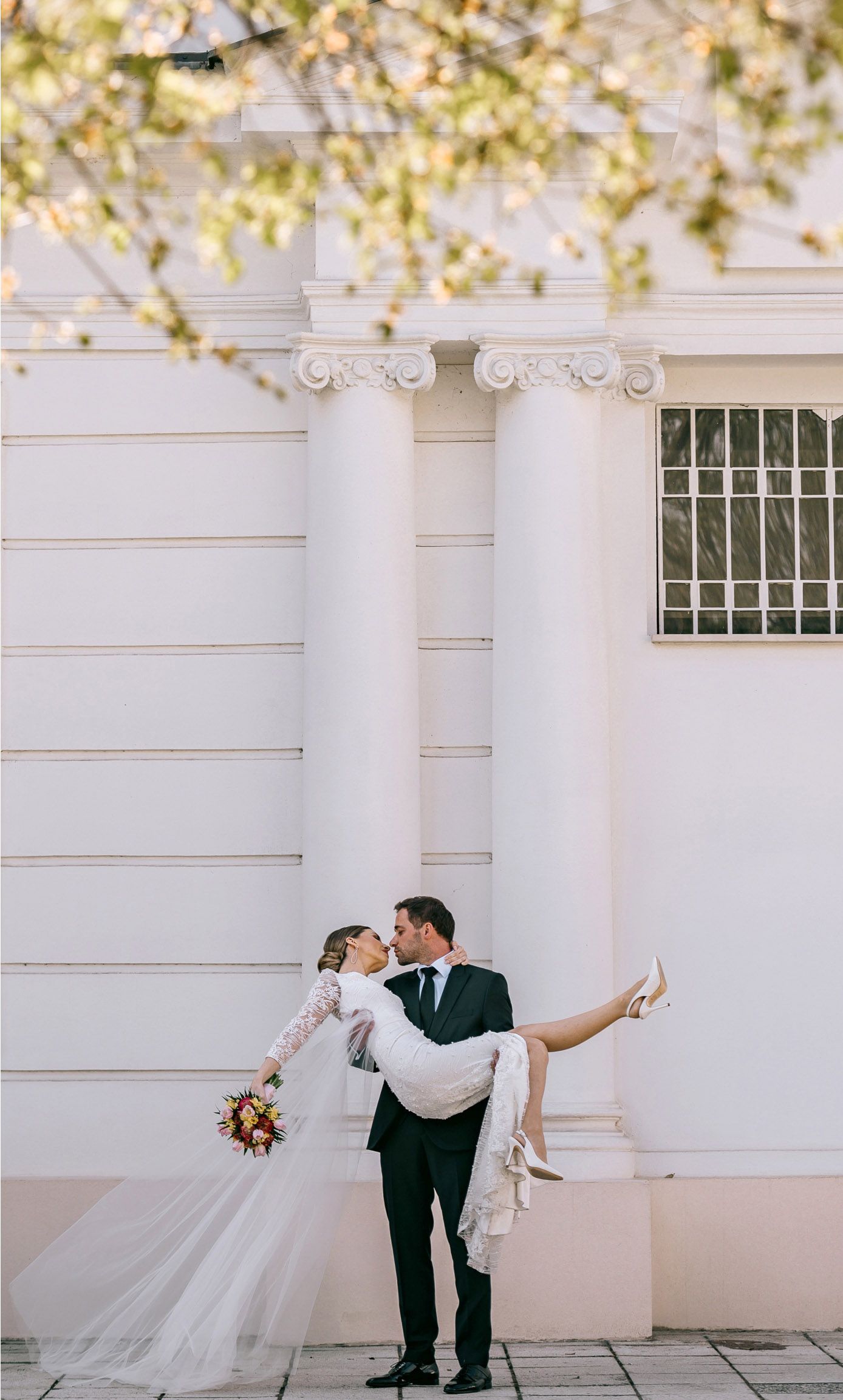 Elegant wedding portrait in front of white columns with bride being carried in playful pose.