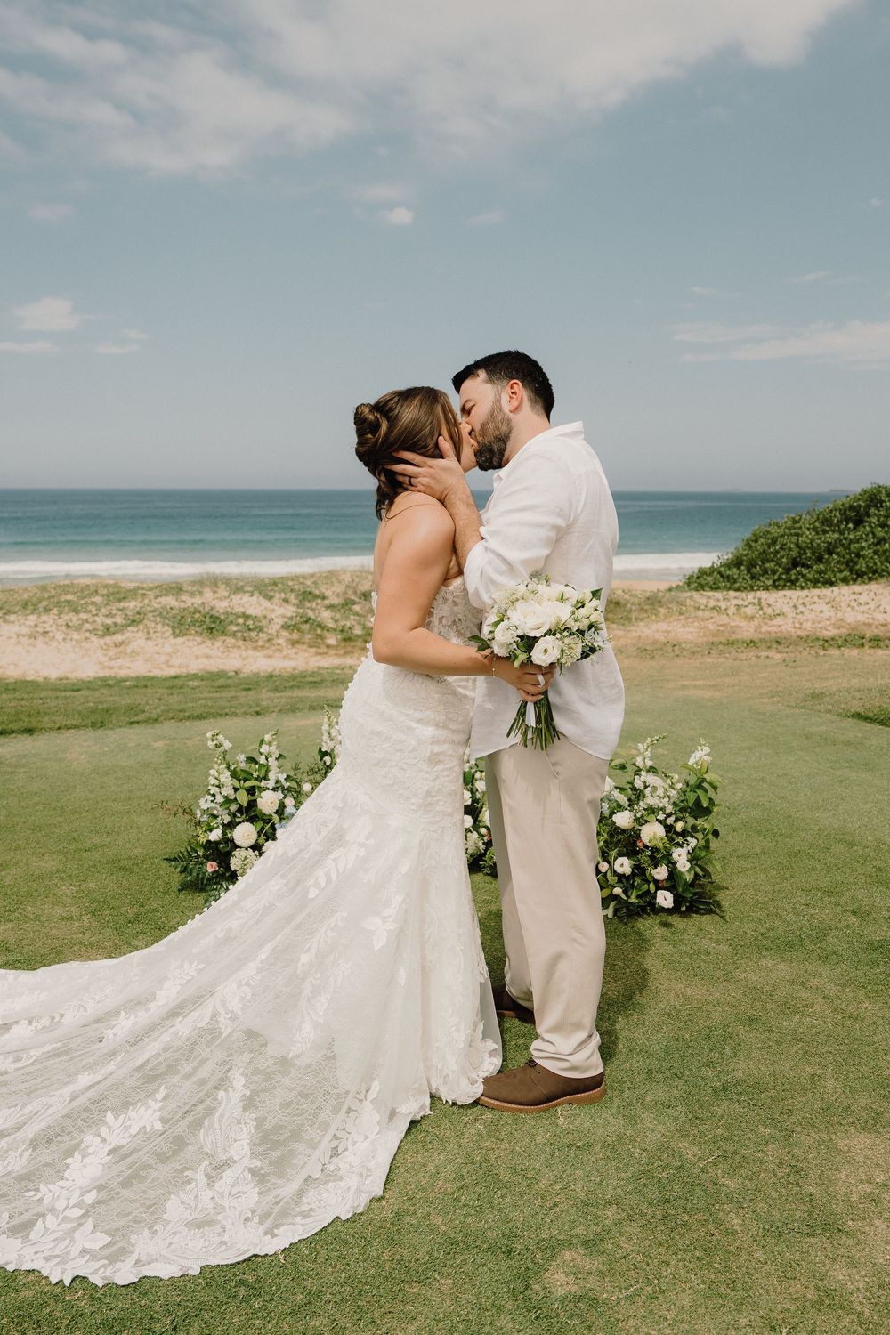 A romantic coastal wedding scene with a couple embracing on a grassy cliff overlooking the ocean.