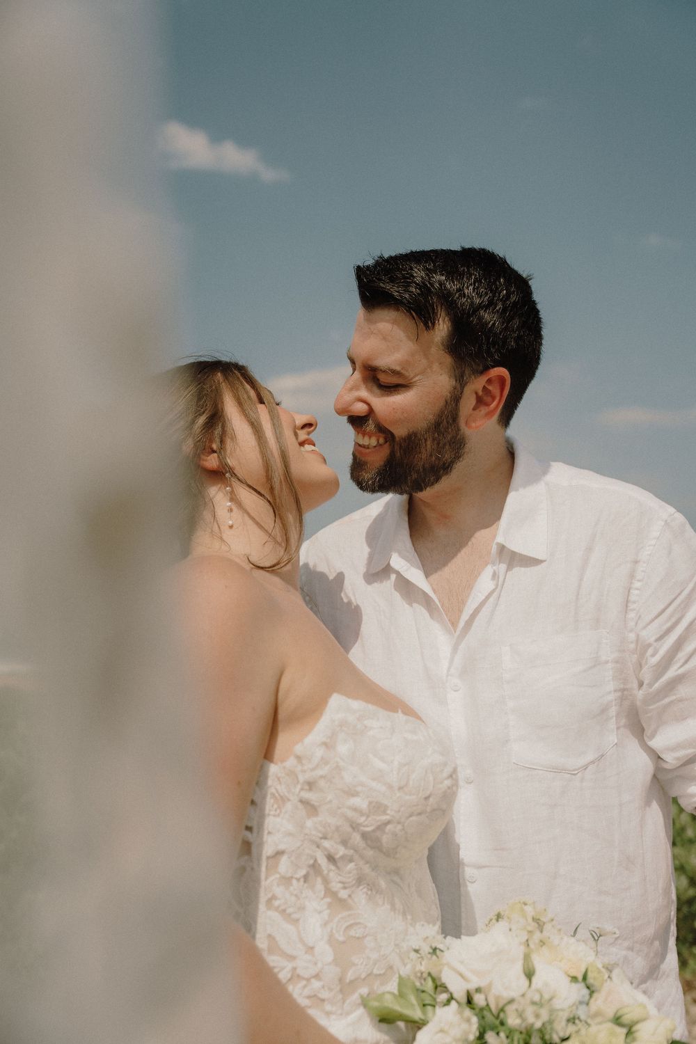 A romantic couple in white wedding attire share an intimate moment against a soft blue sky background.