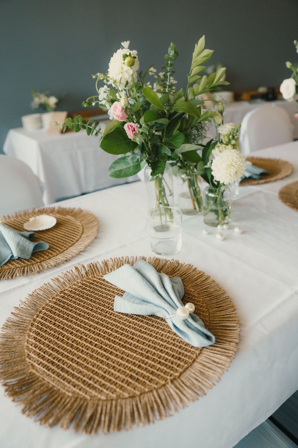 Wedding reception table decor featuring natural woven placemats, blue napkins and fresh flower arrangements.