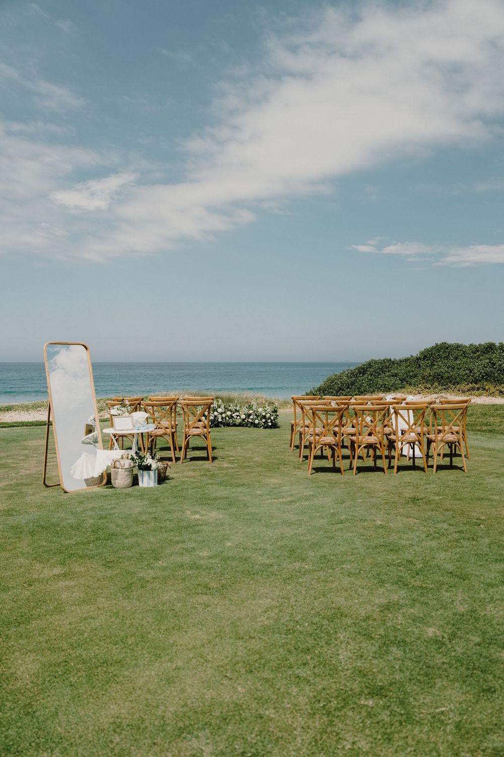 Outdoor wedding ceremony setup with wooden chairs and white decor elements on a grassy oceanfront cliff under blue skies.