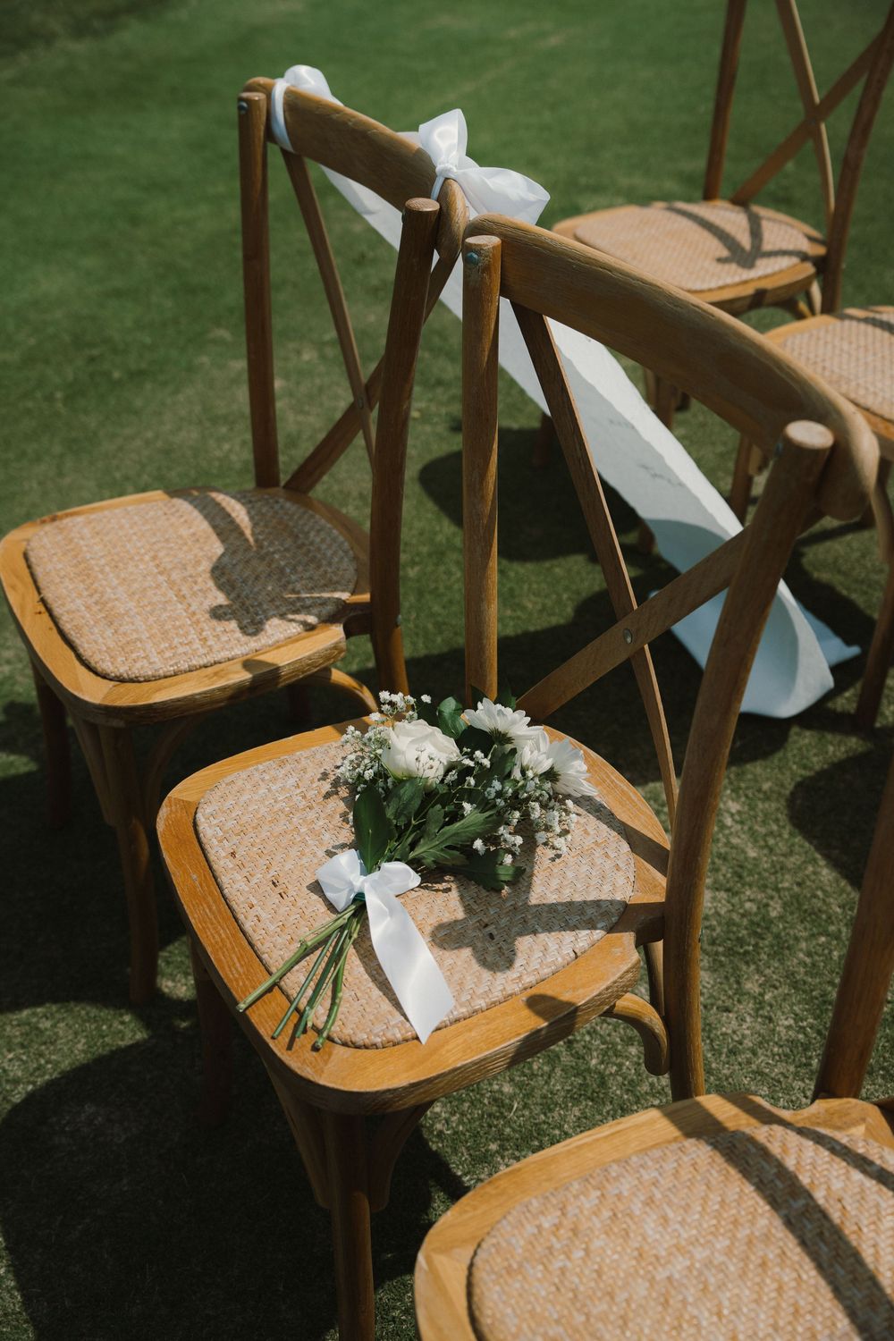 A series of wooden cross-back chairs decorated with white ribbons and small floral arrangements on a grassy lawn.