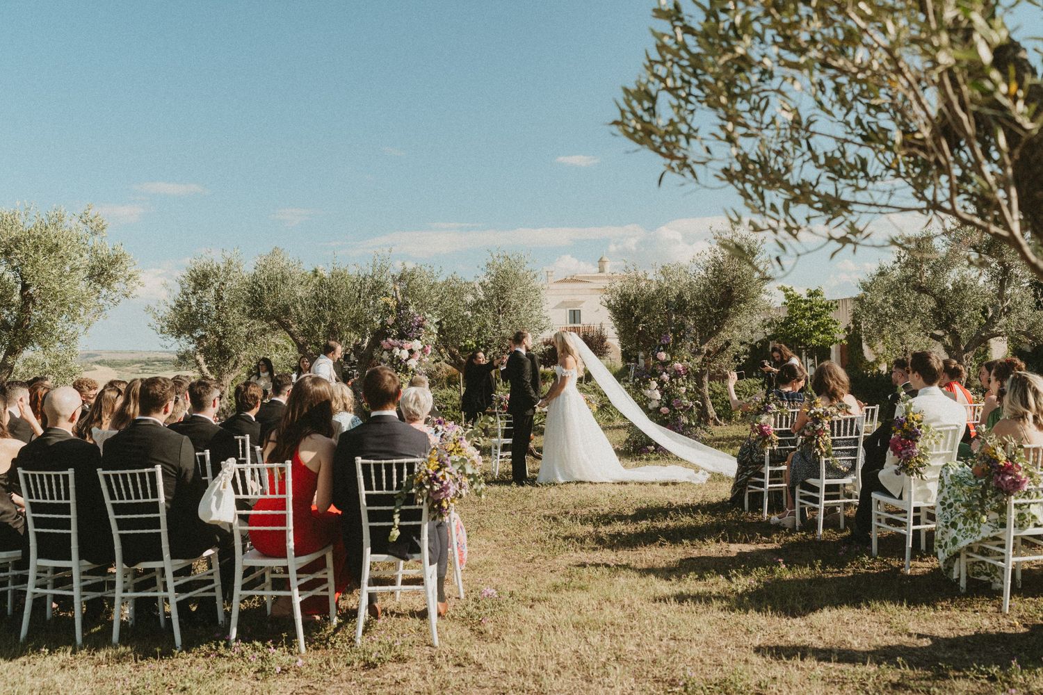 Wedding guests cheer and celebrate as newlyweds walk down an aisle lined with white chairs in an outdoor garden setting.