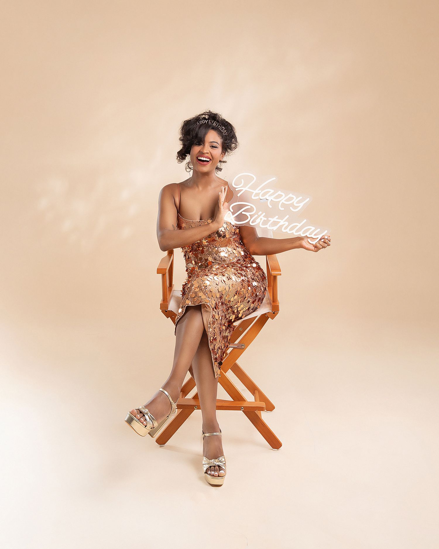 Model in floral dress poses playfully in wooden director's chair against beige background in multiple poses.