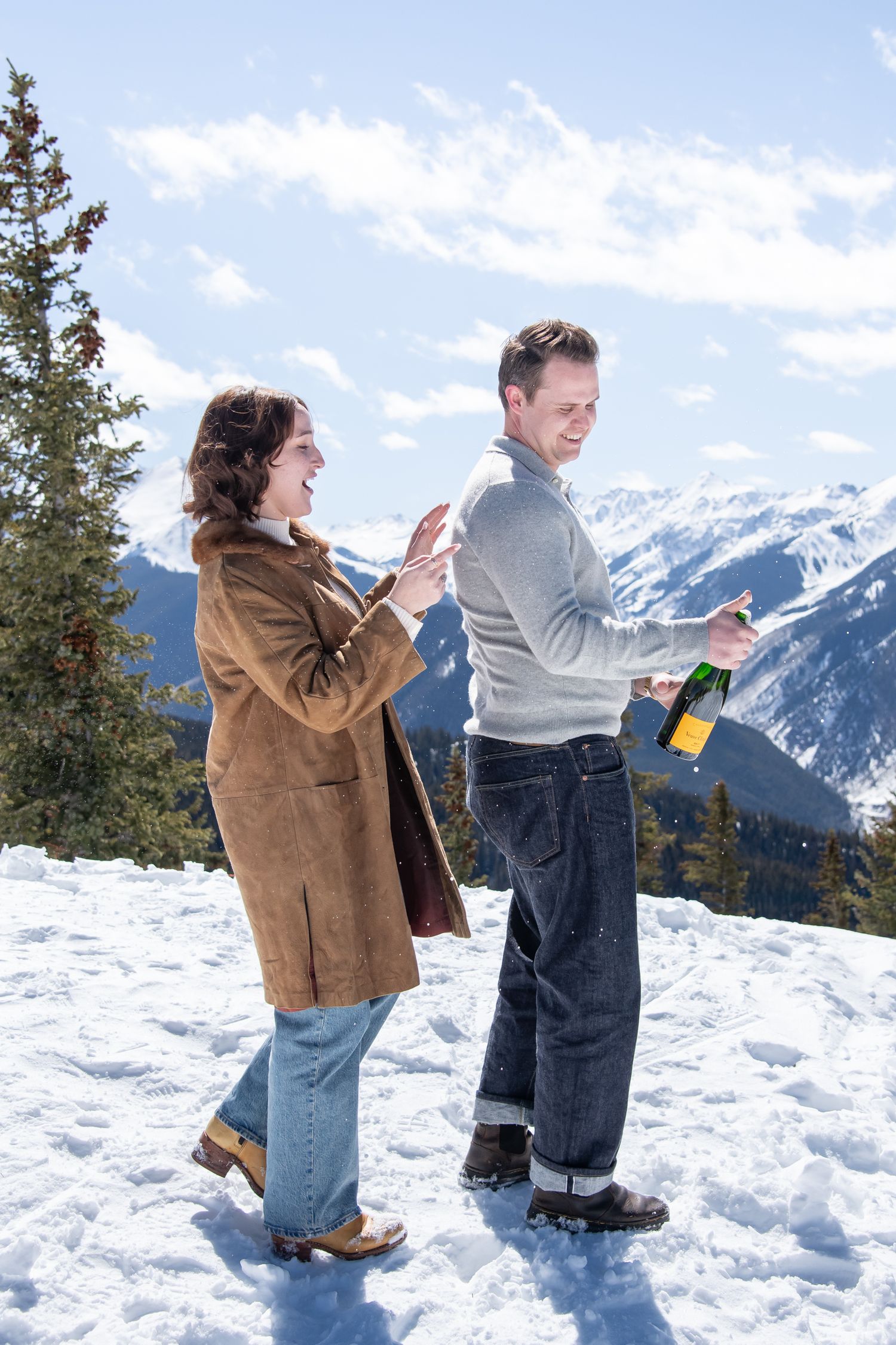 A newly engaged couple pops a bottle of champagne on top of Aspen mountain during a winter engagement photo session.