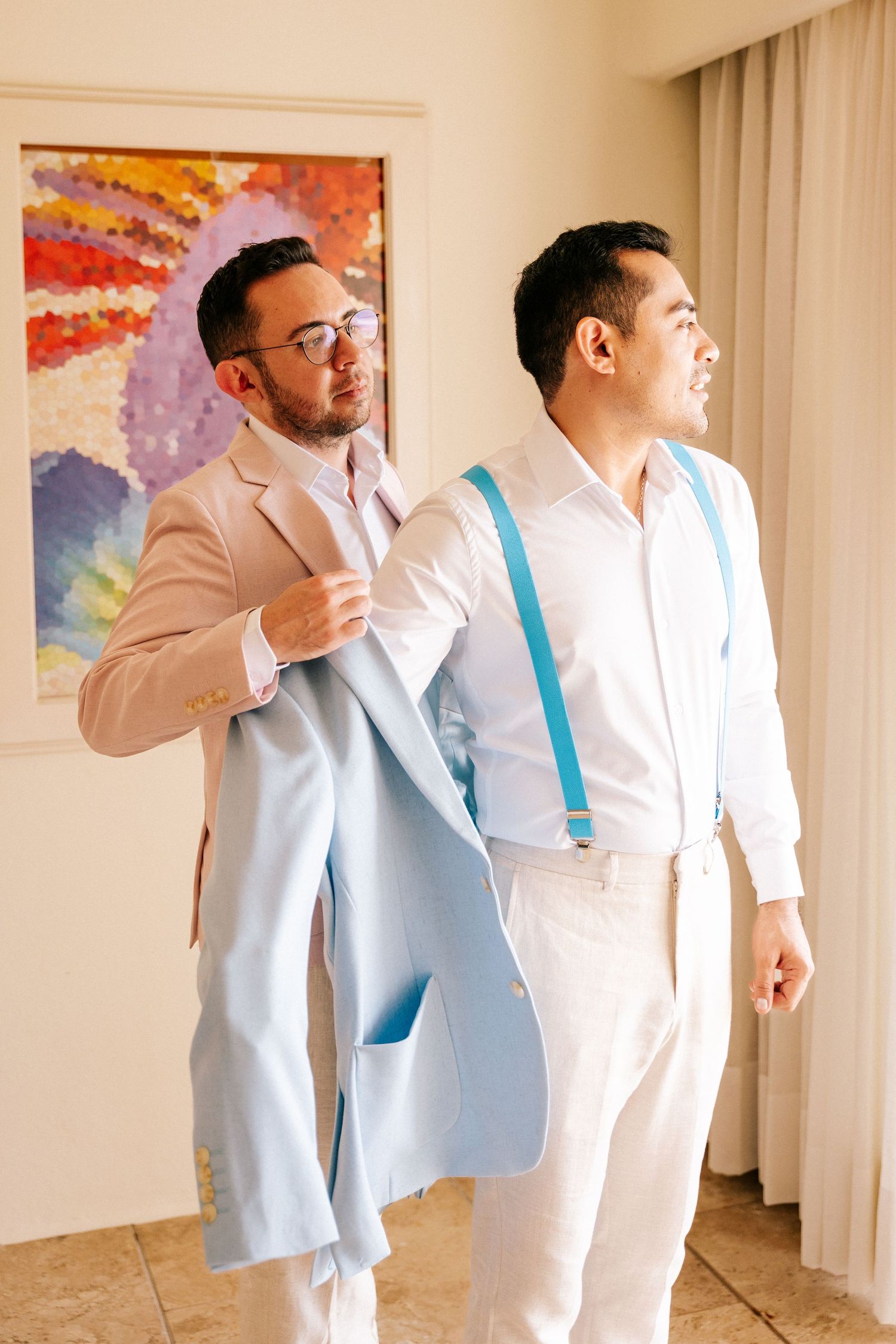 Groomsmen in light colored suits with turquoise accents pose together in an indoor setting with colorful artwork.