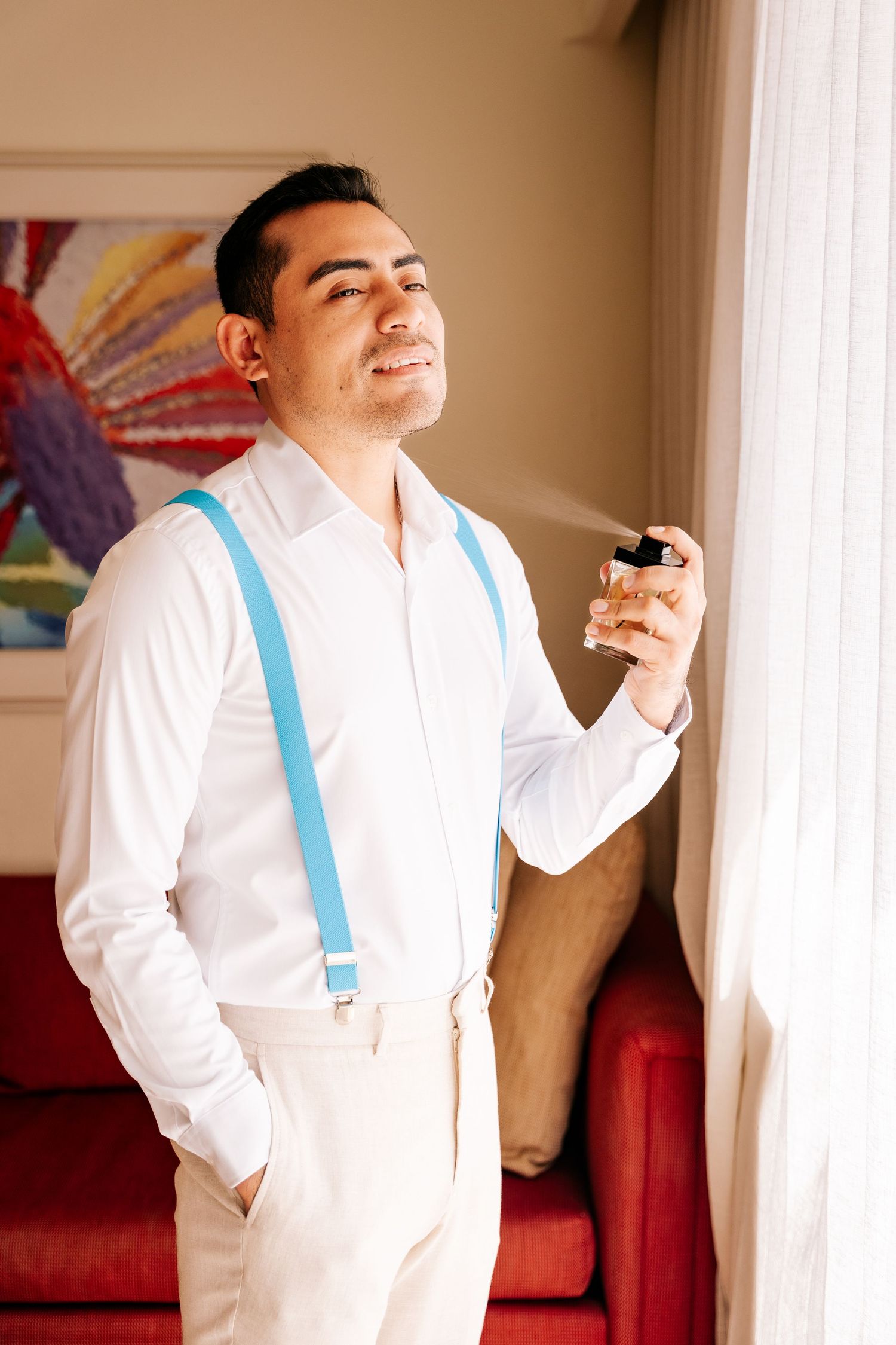 A person in a white dress shirt and light blue suspenders stands near a window with a joyful expression.