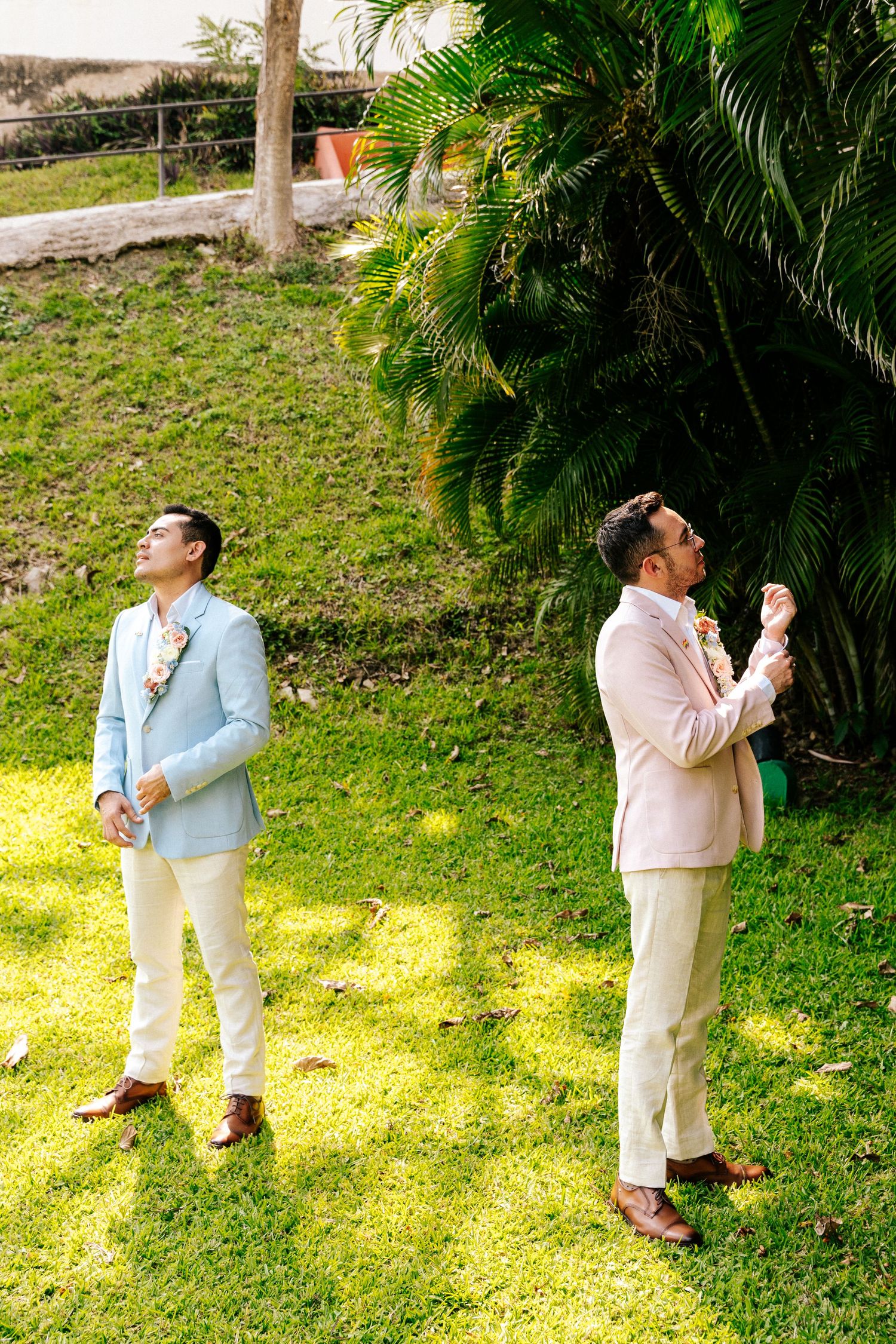 Two people in pastel suits stand back-to-back on a grassy lawn with tropical palm trees in the background.