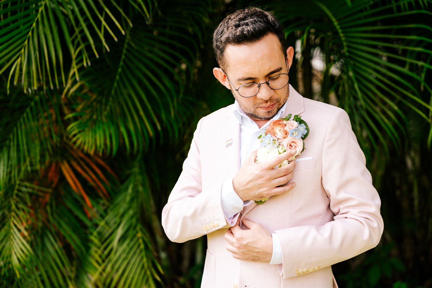 Person in a white suit jacket removes and eats a rainbow pride flag pin against a tropical palm background.