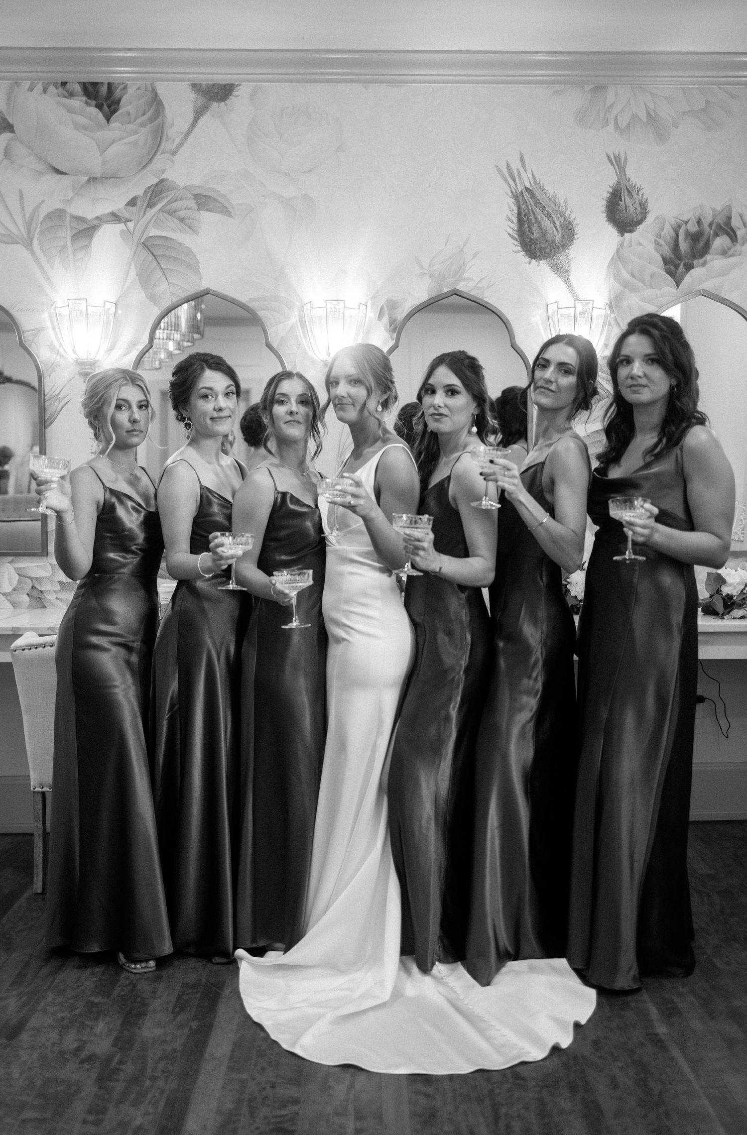 Bride in white gown poses with six bridesmaids in dark satin dresses holding champagne glasses.