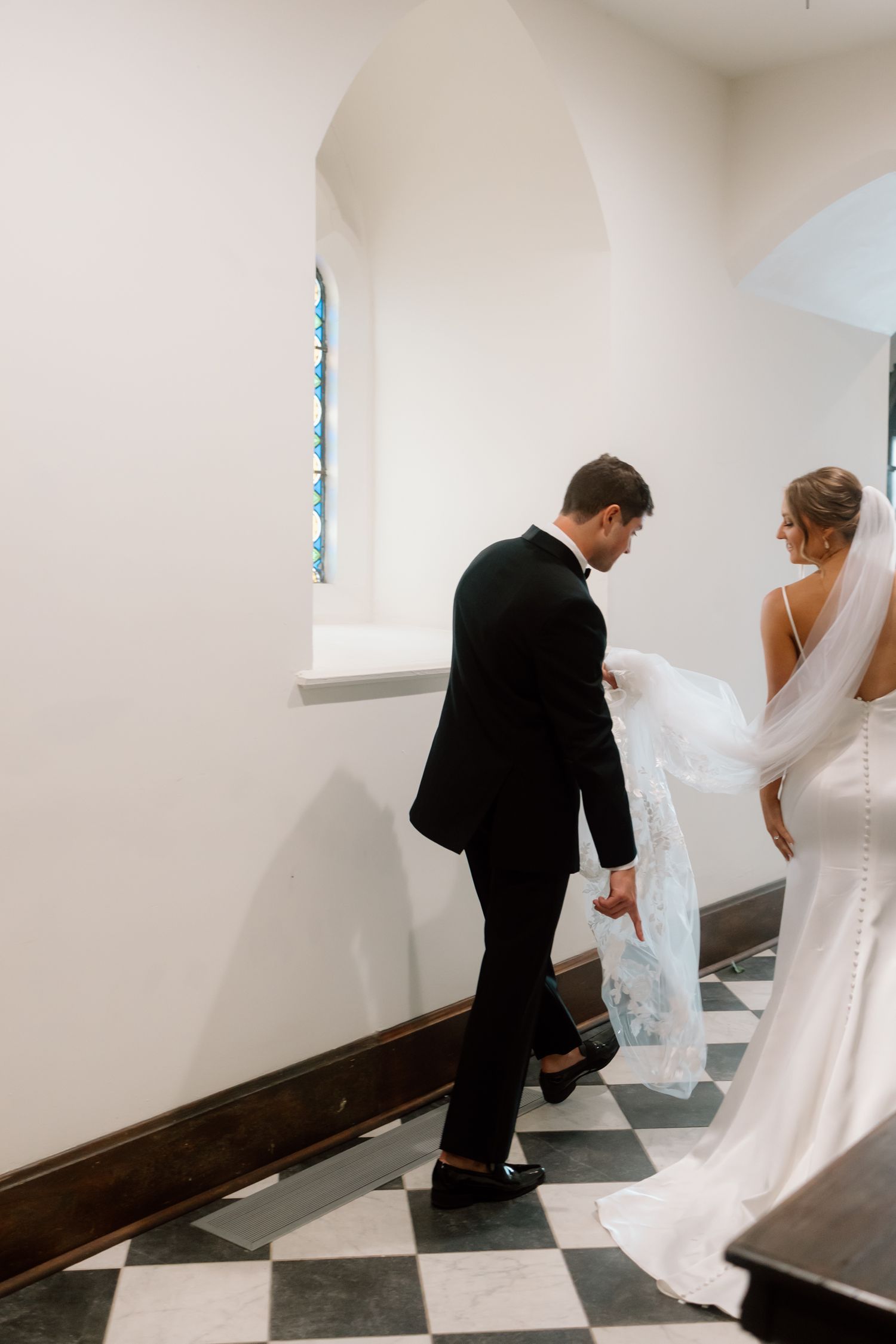 Bride and groom walking through church hallway, groom carrying wedding dress train on checkered floor.