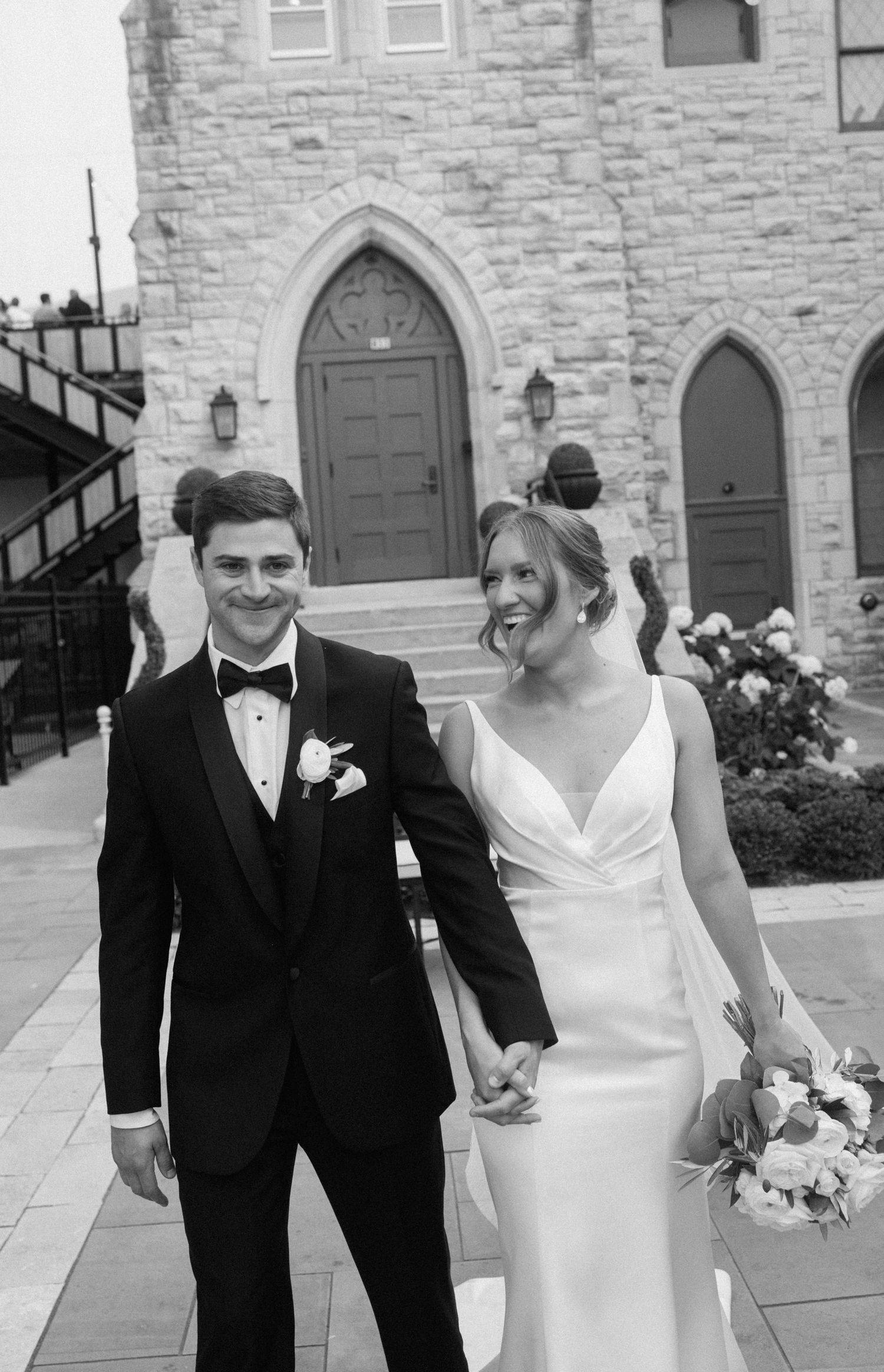 Black and white photo of smiling bride and groom holding hands outside a stone church after their wedding ceremony.