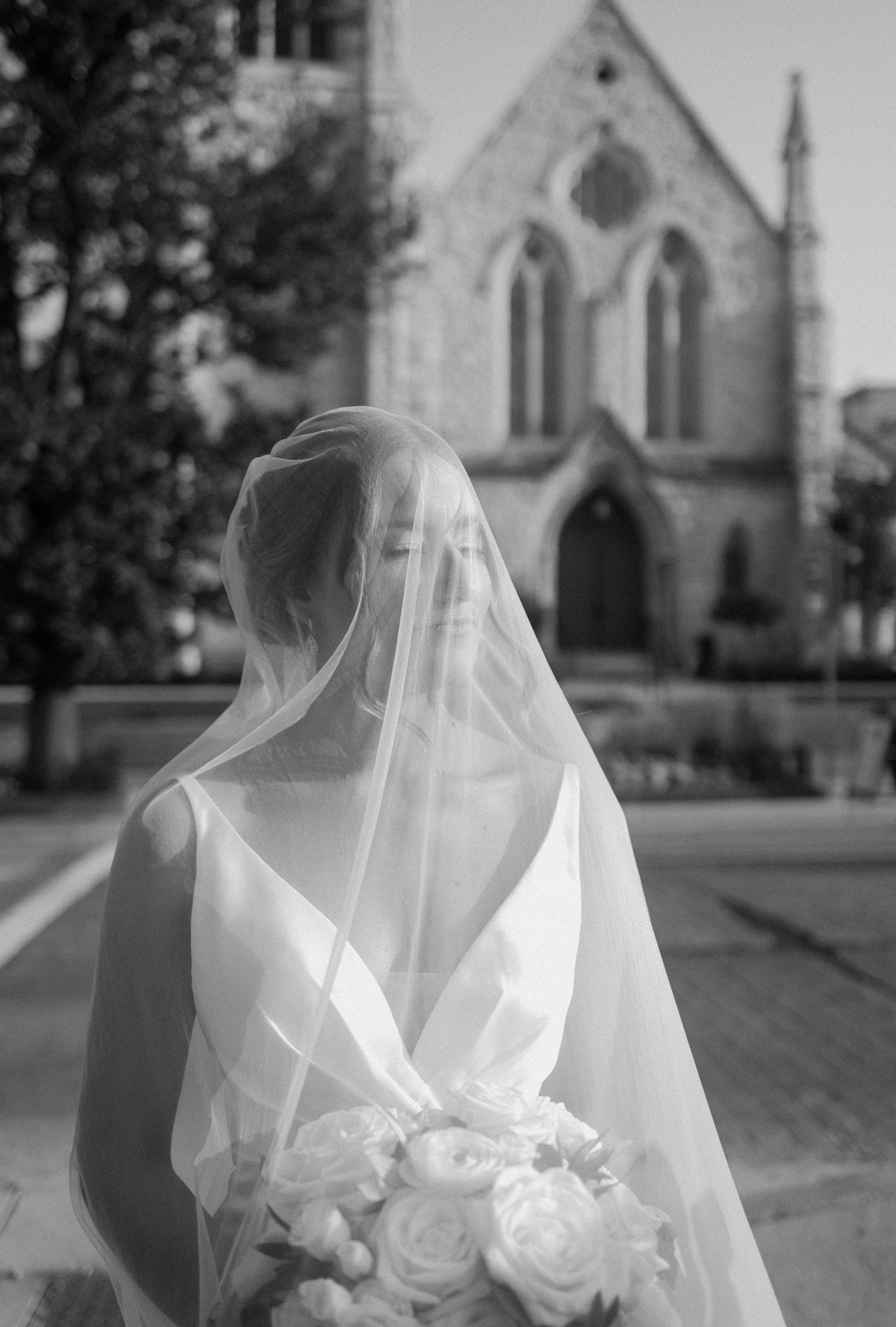 Bride in white gown and veil holding bouquet outside stone church, black and white wedding photo.