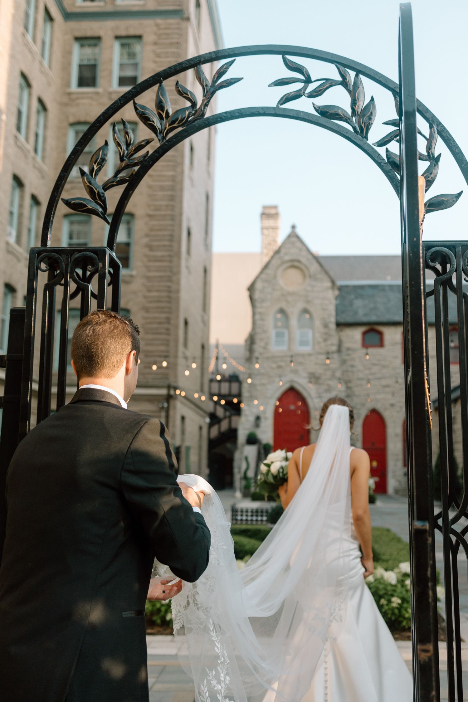 Bride and groom walking through ornate iron arch toward stone church with red doors and string lights.