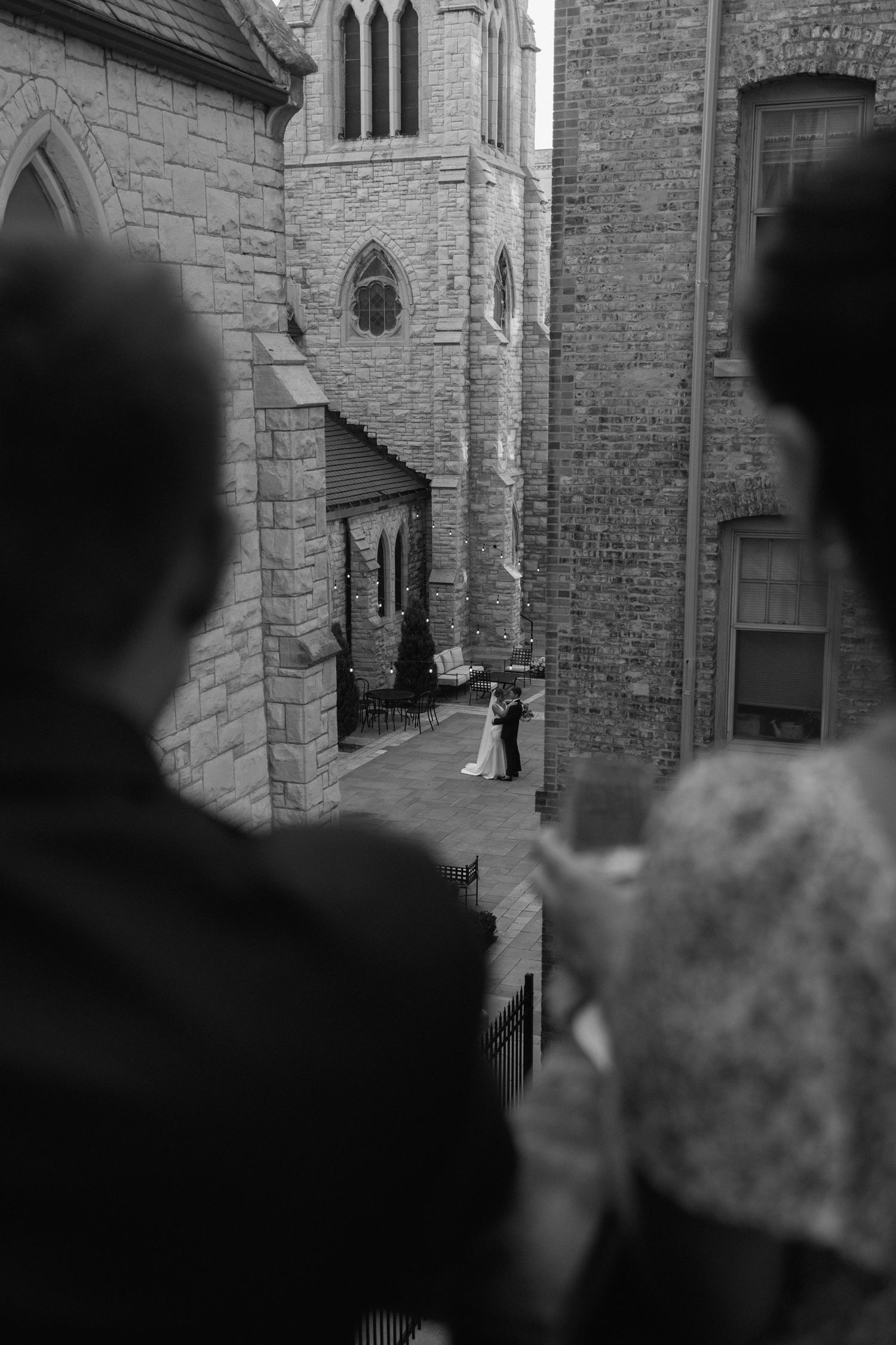 Aerial black and white view of bride and groom dancing outside historic stone church with onlookers.