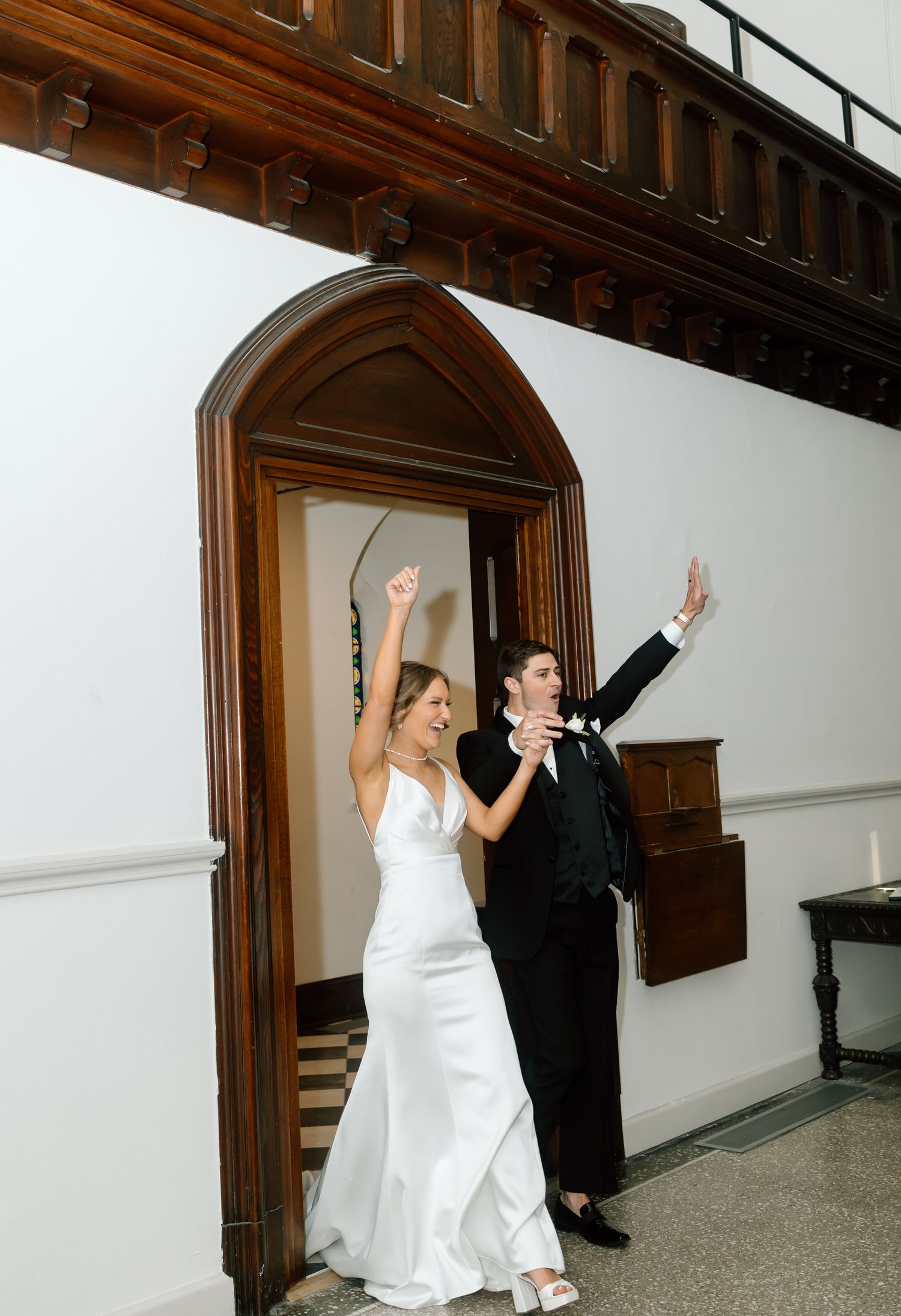 Joyful bride in white gown and groom in tuxedo raising arms in celebration as they exit through ornate wooden church doors.