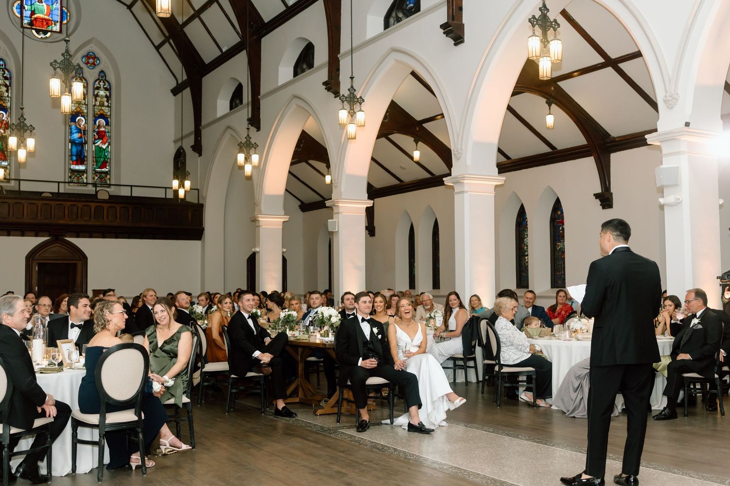 Elegant wedding reception in a historic church venue with arched ceilings, stained glass windows, and guests seated at round tables.