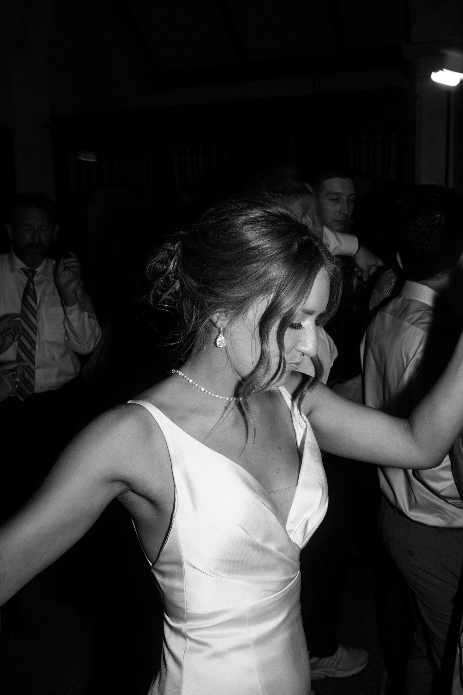 Bride in white satin gown dancing joyfully at wedding reception, black and white photo with guests in background.