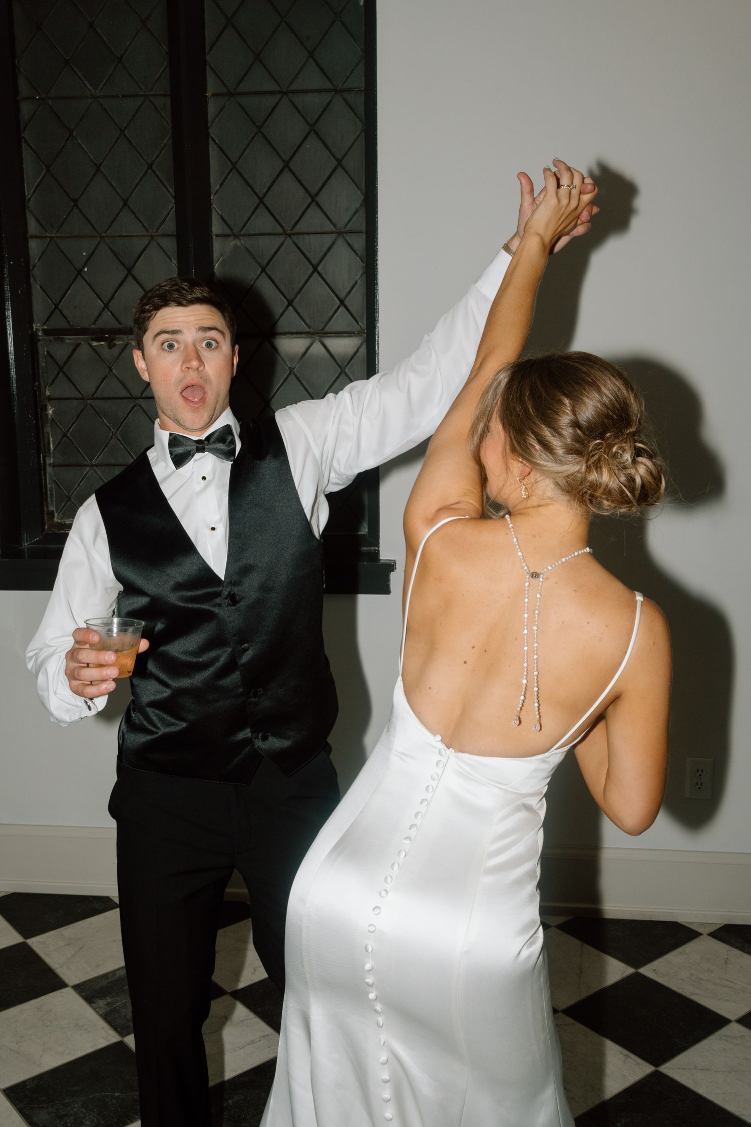 Bride and groom dancing joyfully at wedding reception, groom in black tuxedo holding drink, bride in elegant backless white gown.