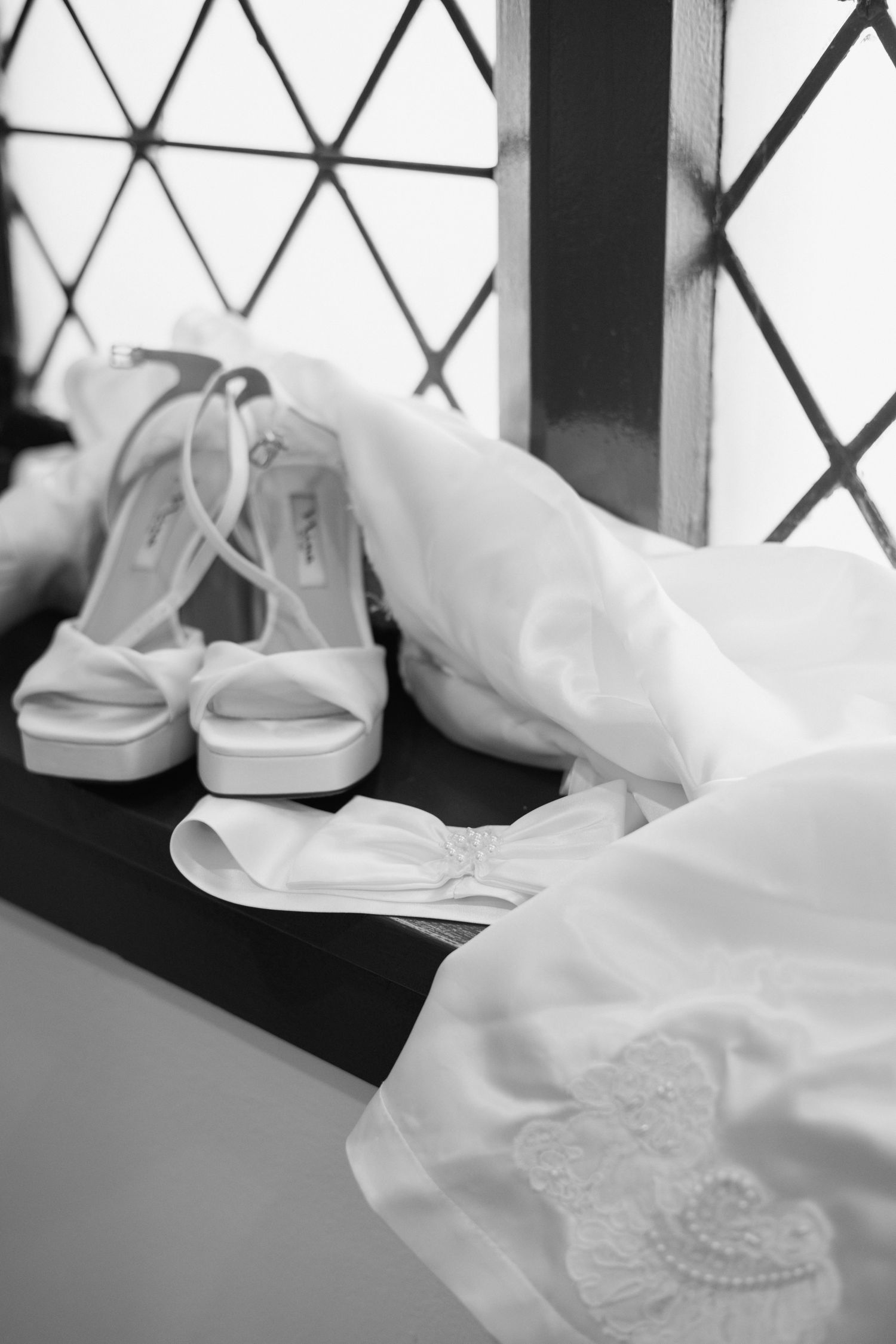 White bridal heels and wedding dress accessories arranged on a windowsill in black and white photography.