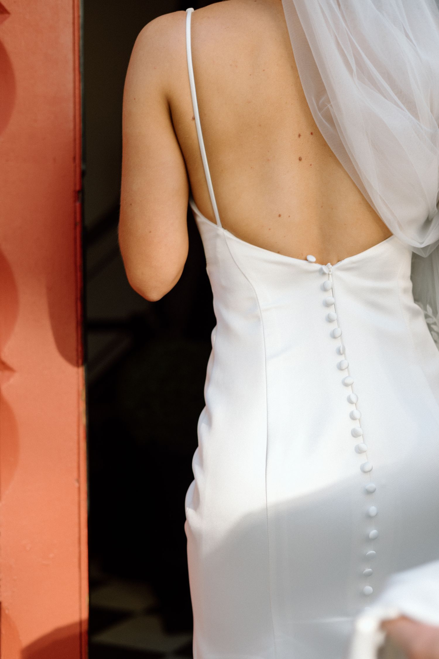 Close-up back view of bride in sleek white satin gown with delicate button detail and sheer veil.