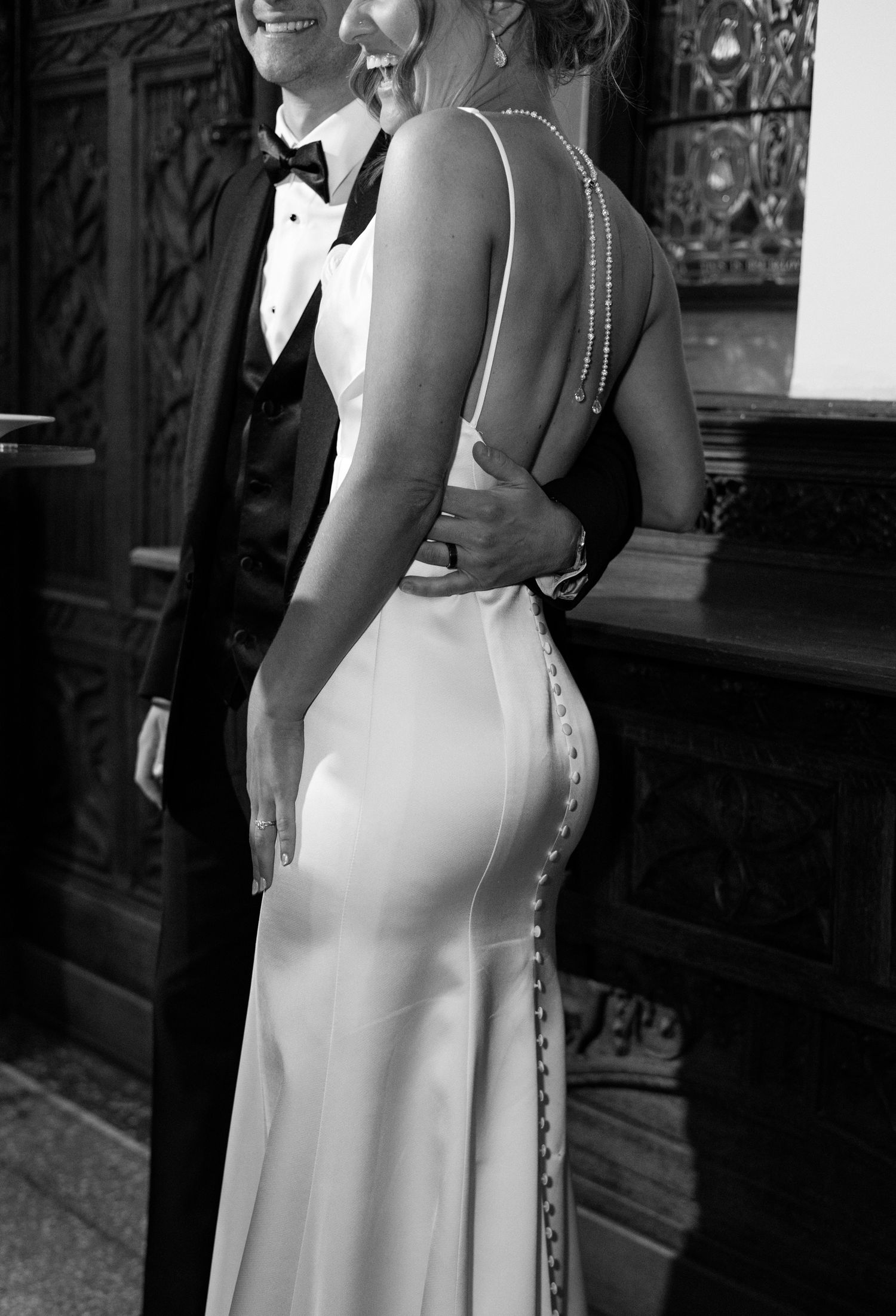 Black and white photo of bride in backless satin gown with crystal details embraced by groom in tuxedo.