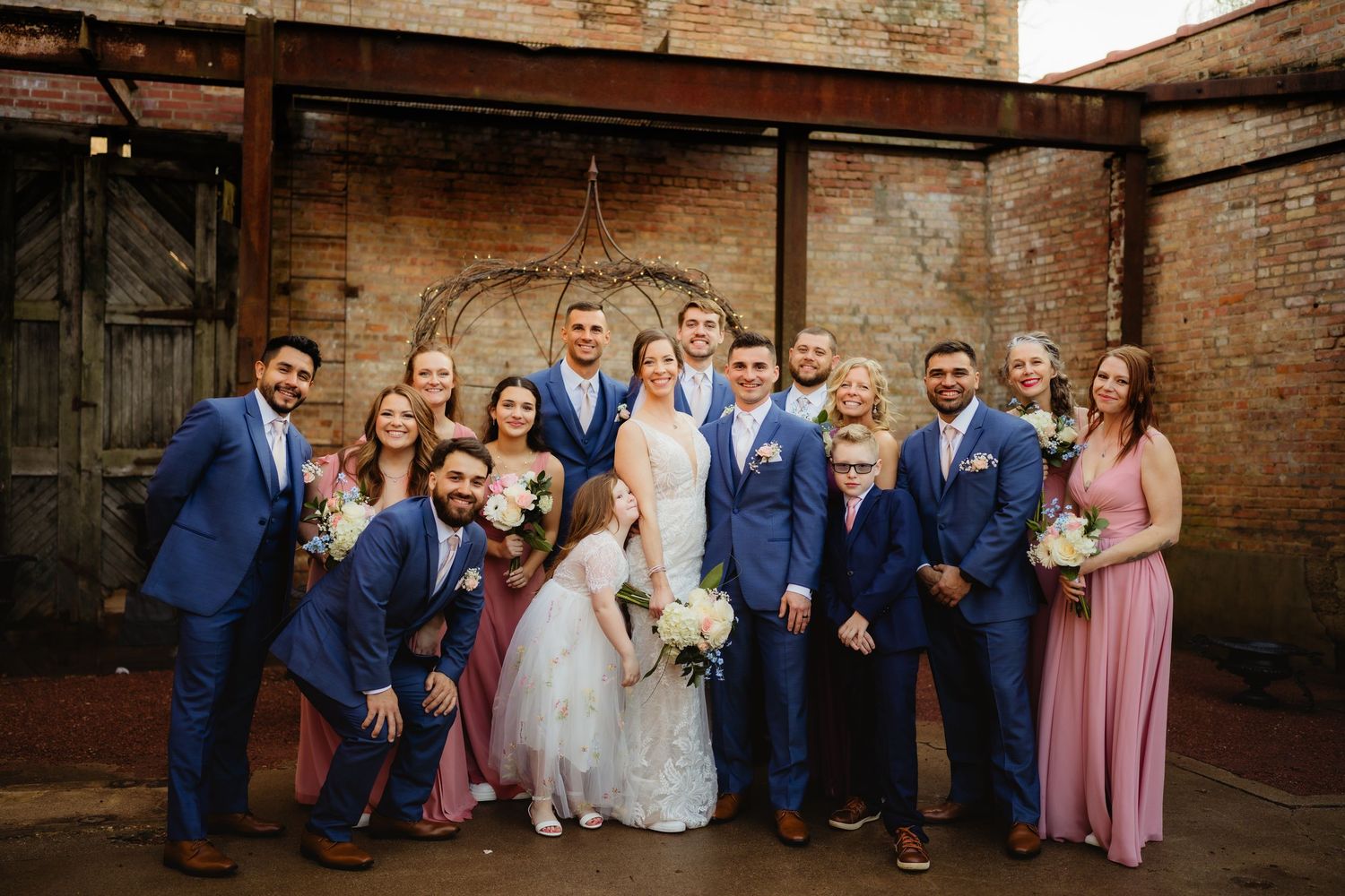 Wedding party in blue suits and pink dresses posing together outdoors at a rustic brick venue.
