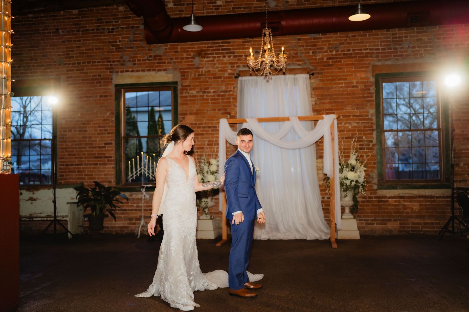 Bride and groom share first dance in rustic brick venue with chandelier and floral arch backdrop.