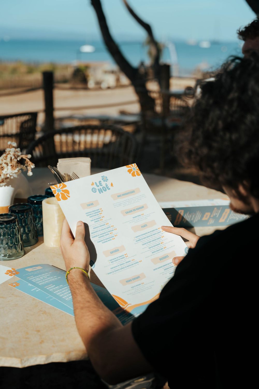Homme lisant un menu de restaurant bleu et orange dans un café en plein air au bord de la plage avec vue sur l'océan.