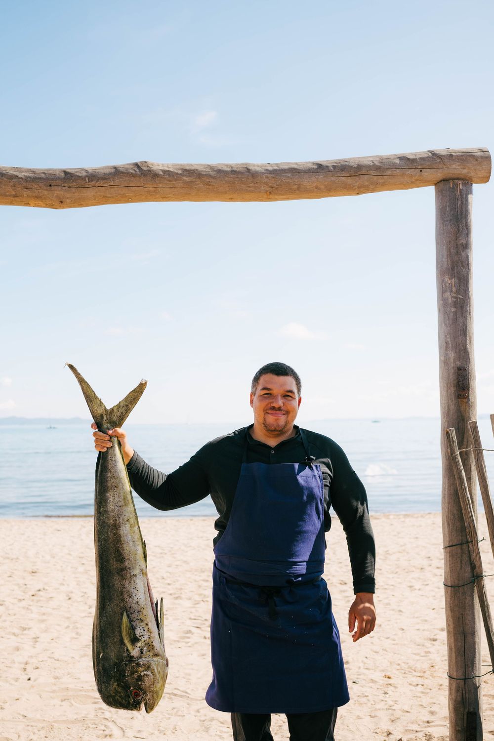 Le chef en tablier bleu tient de gros poissons frais sur une plage de sable avec une toile de fond océanique.