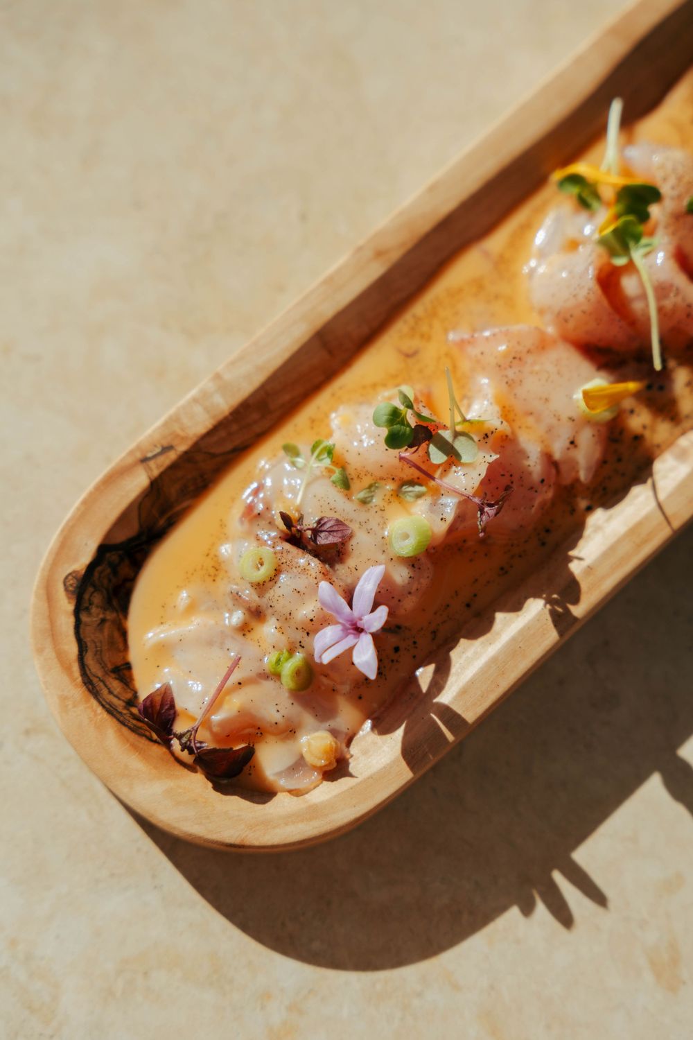 Élégant plat de sashimi garni de fleurs comestibles et de microgreens, servi dans un plateau en bois rustique avec une sauce crémeuse.