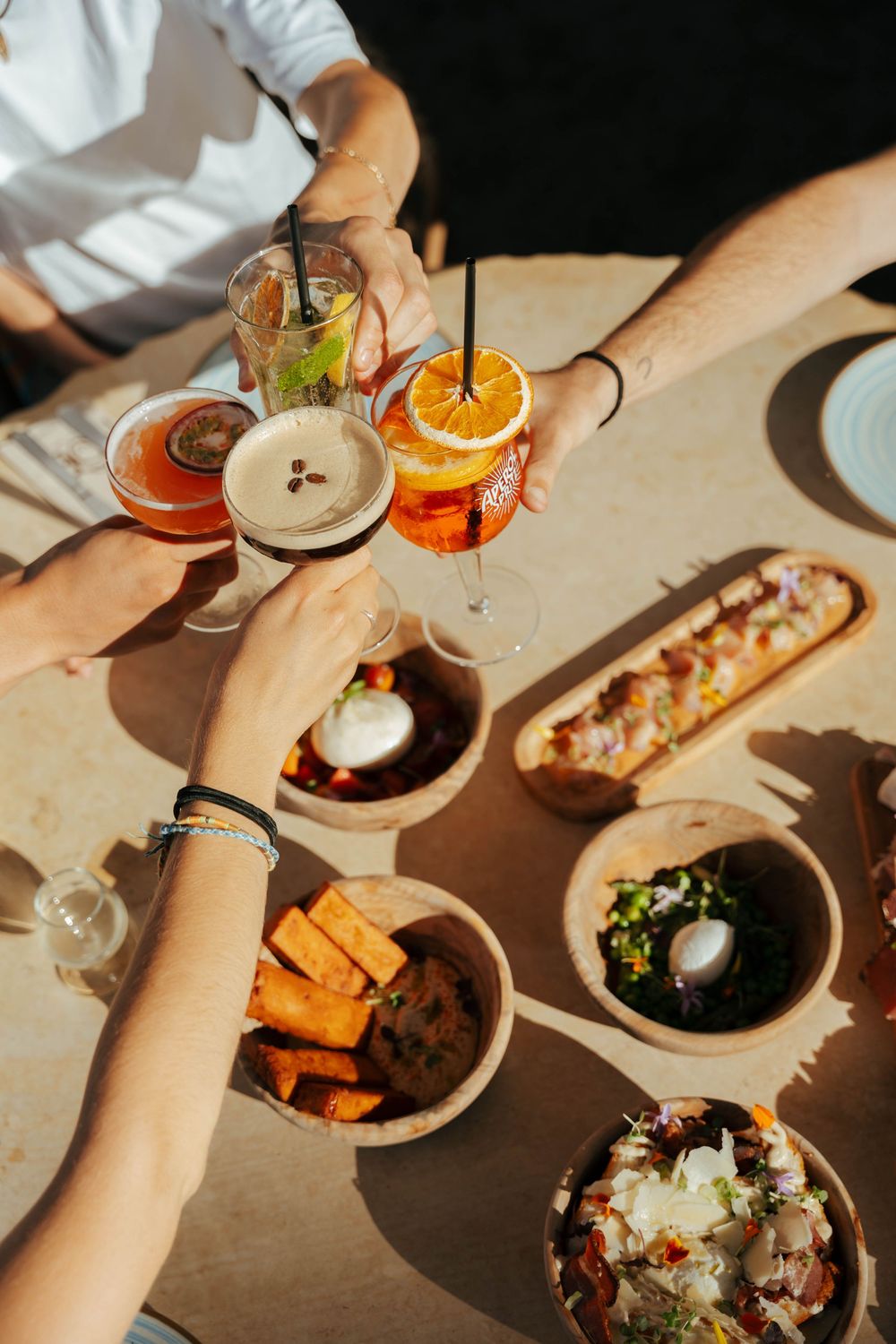 Friends clinking cocktails over bowls of food and appetizers on a sunny outdoor dining table.