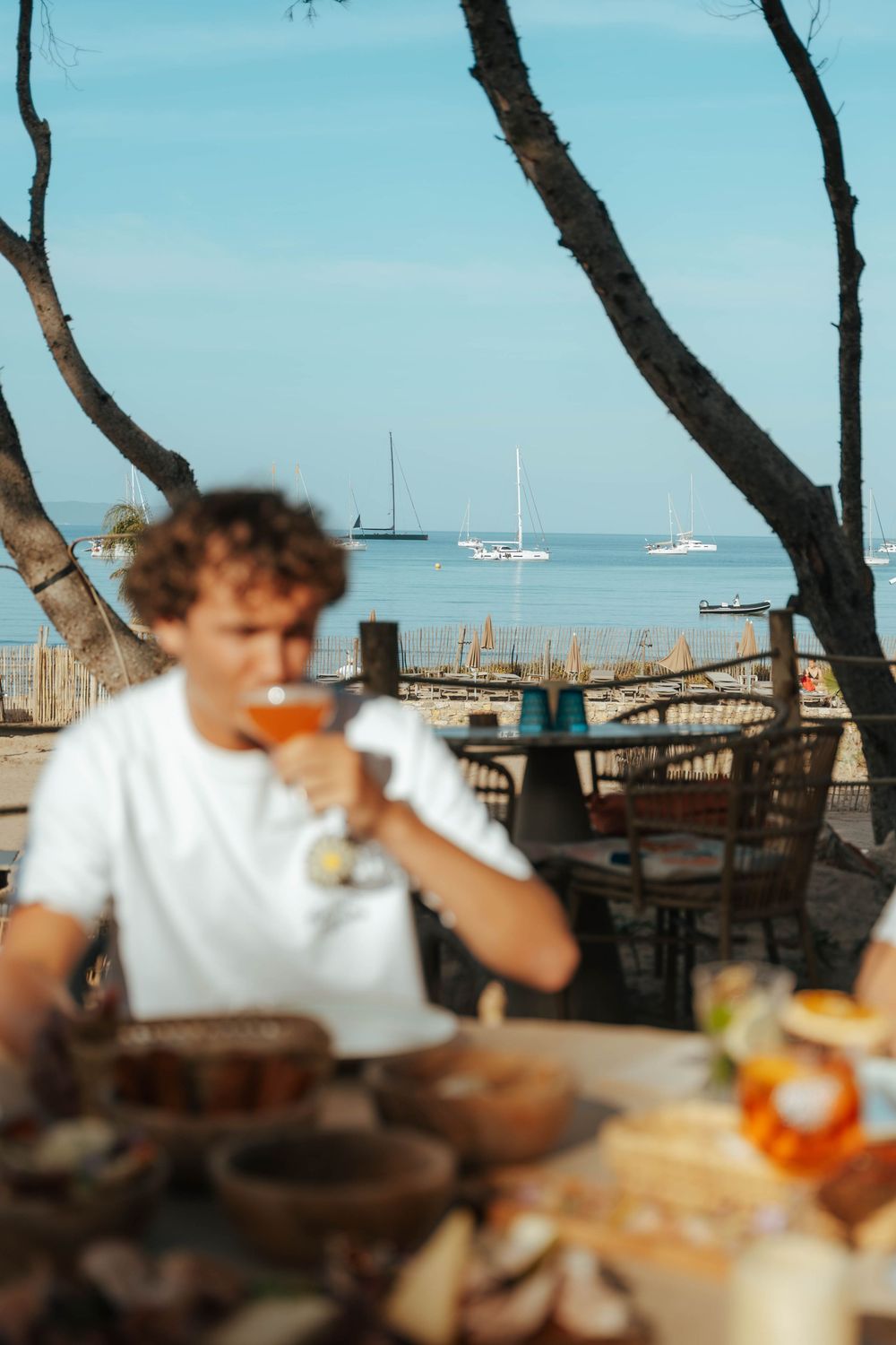 Jeune homme sirotant du vin rosé à une table de restaurant au bord de la plage avec des voiliers et une mer calme en arrière-plan.