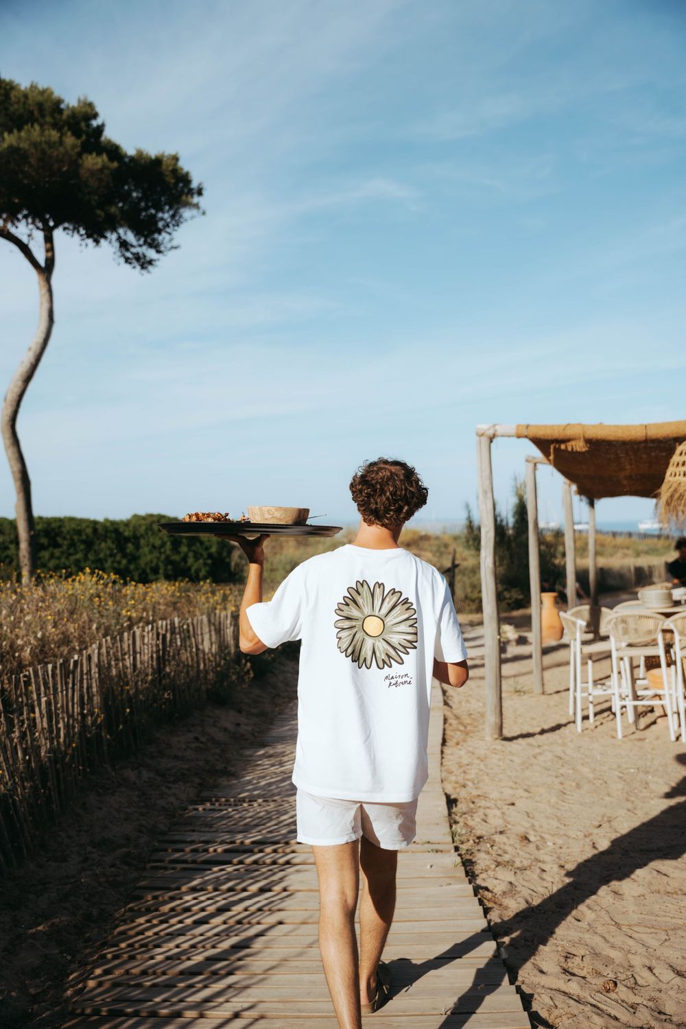 Homme en t-shirt graphique blanc portant un plateau sur un chemin de promenade de plage ensoleillée.