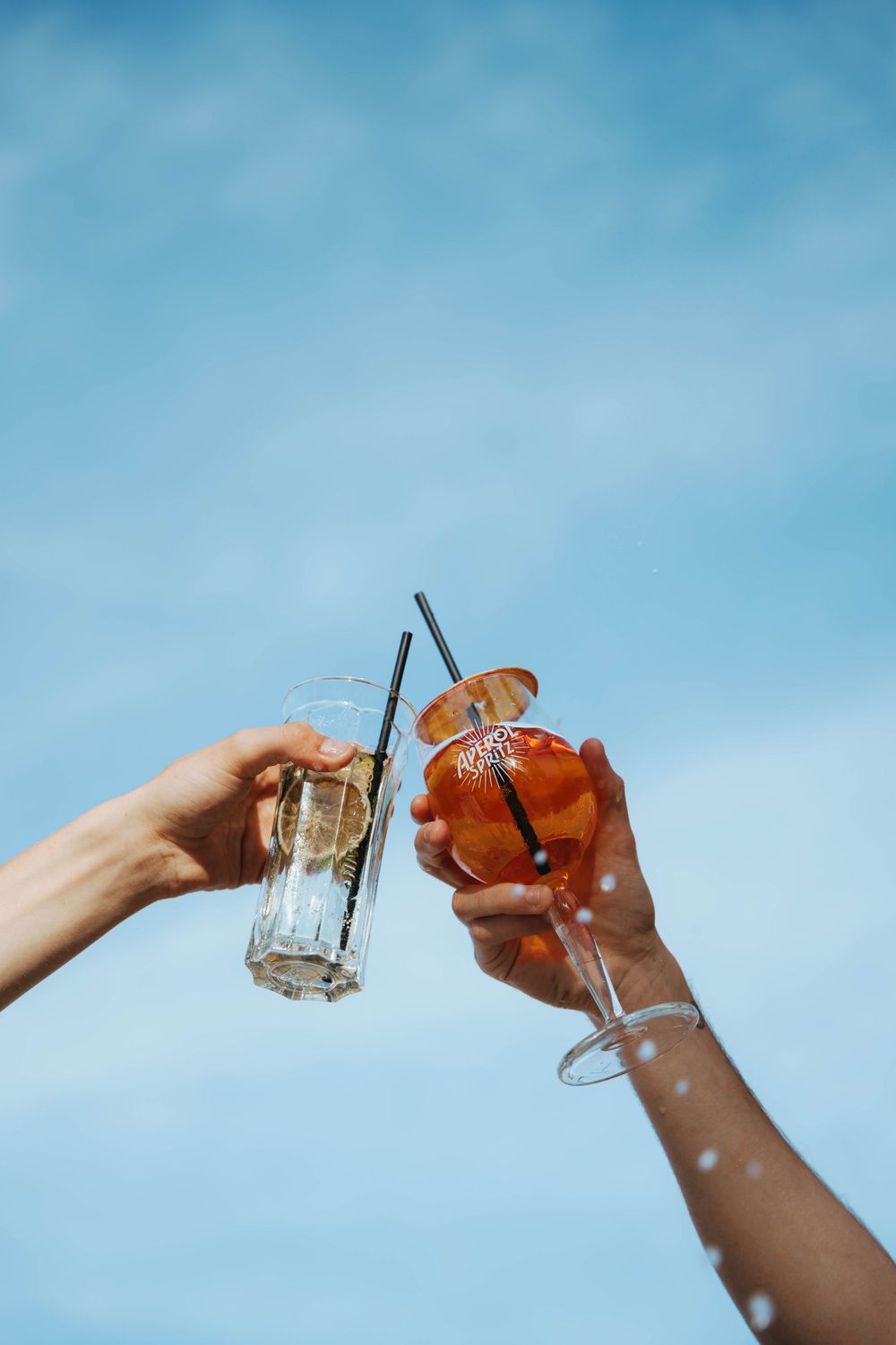Deux mains faisant tinter des verres à cocktail contre un ciel bleu, trinquant avec une boisson claire et un Aperol Spritz orange.