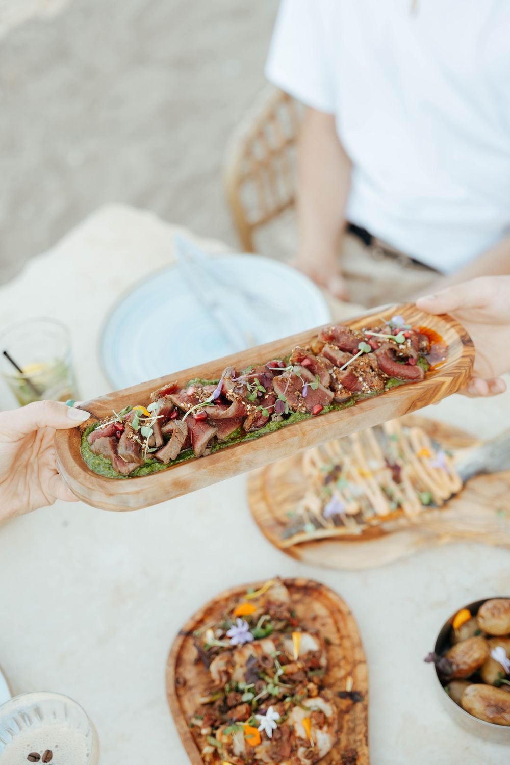 Hands passing a wooden platter of seared tuna with microgreens at a beach dining table with flatbreads.