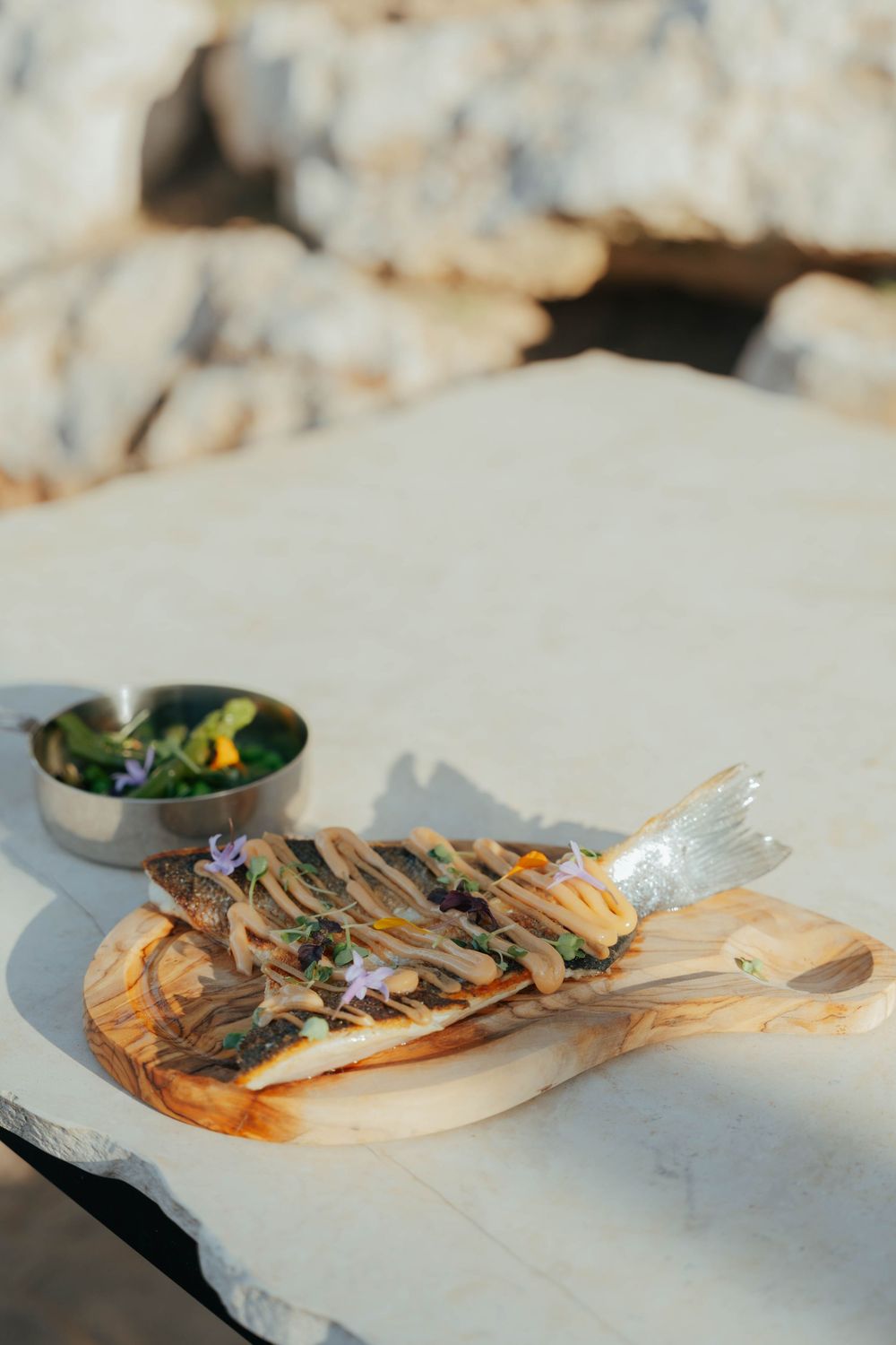Poisson grillé servi sur une planche de bois avec des fleurs comestibles, à côté d'un petit bol de salade sur une surface en marbre.